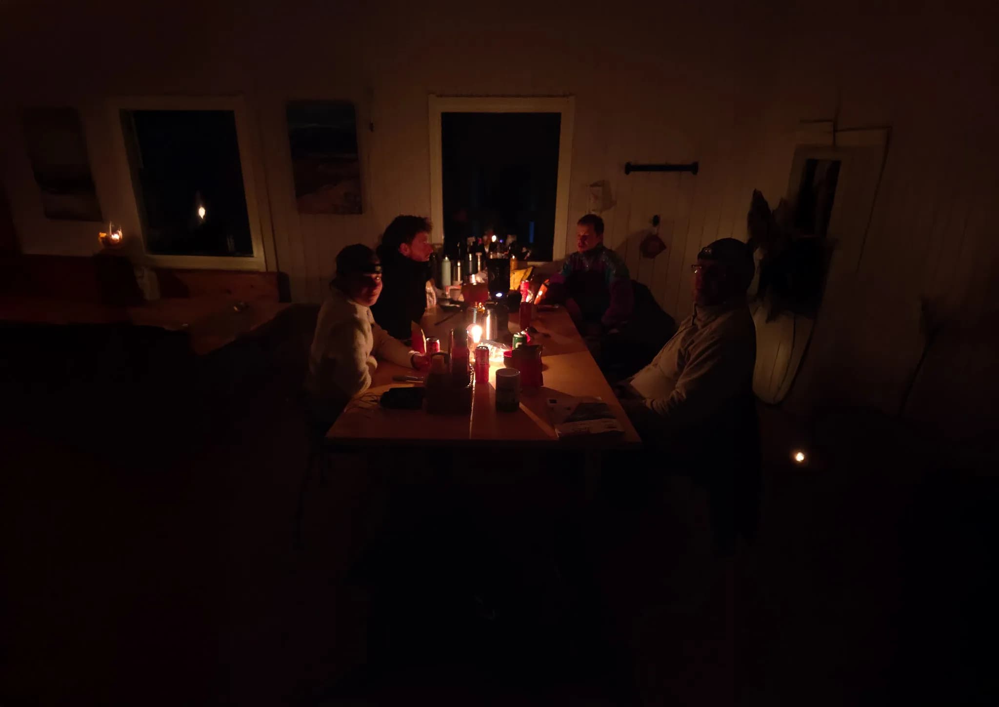 A group of people gathered around a large table in a dimly lit hut, lit mainly by candles and lamps. The atmosphere is warm and intimate, with faces lit up by the light.
