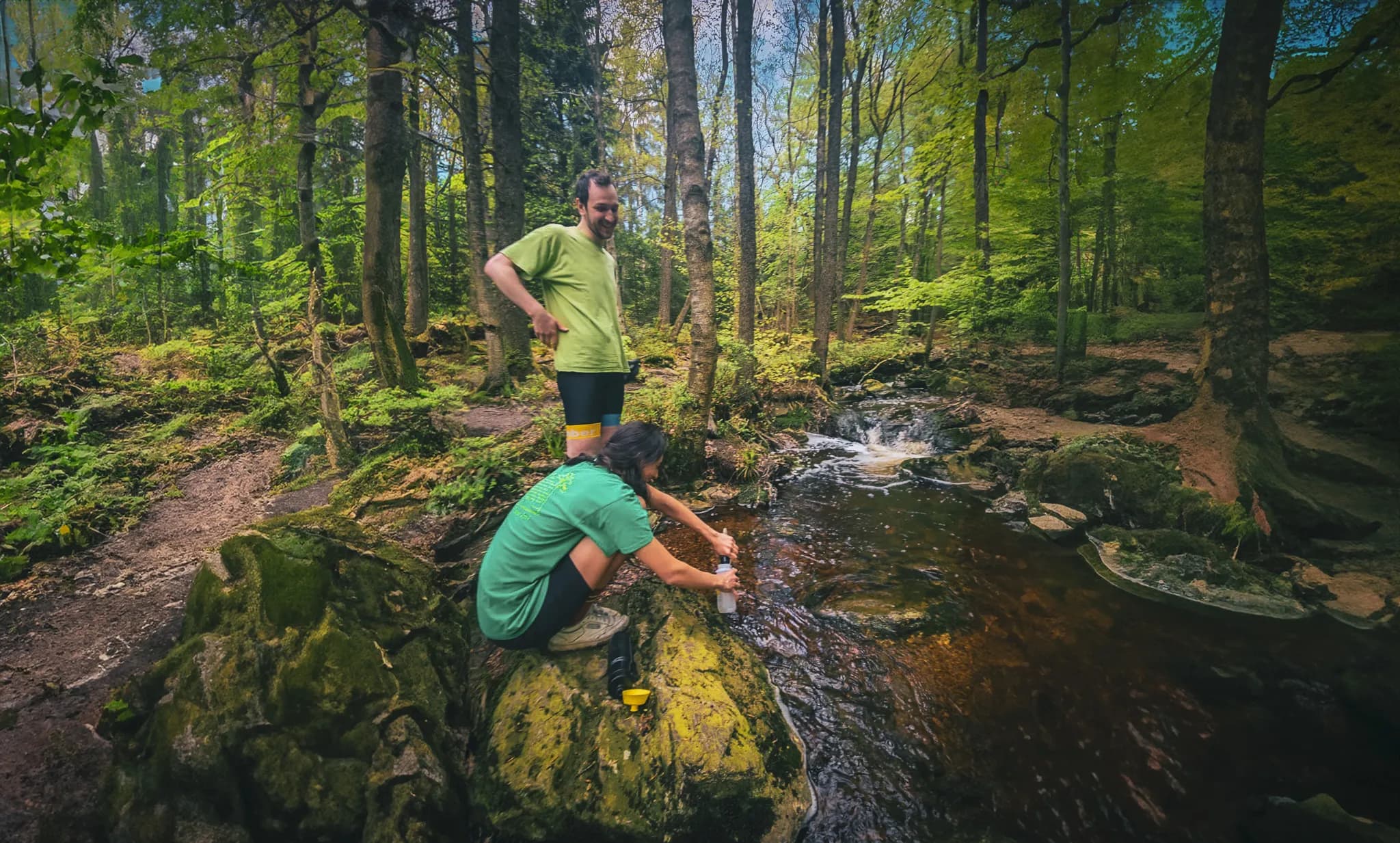Two people are standing by a stream in a green forest. One of them is standing, wearing a green T-shirt and shorts, while the other is crouched on a rock, wearing a green T-shirt and shorts.