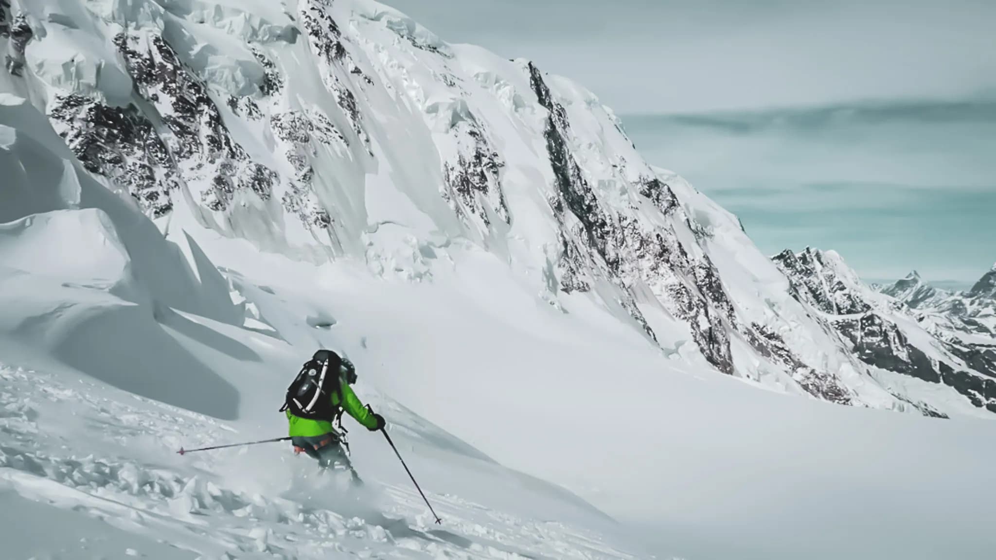 Een skiër daalt af van een smetteloze besneeuwde helling, omringd door majestueuze besneeuwde bergen. De skiër draagt een felgroene jas en een rugzak, terwijl de sneeuwvlokken op de helling nog zichtbaar zijn.