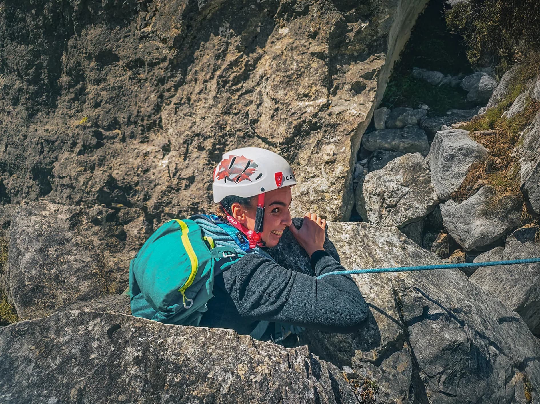A smiling climber, wearing a black jacket and carrying a blue rucksack, climbs a rock face in a natural setting. He is wearing a white safety helmet and holding a blue rope. The light from the