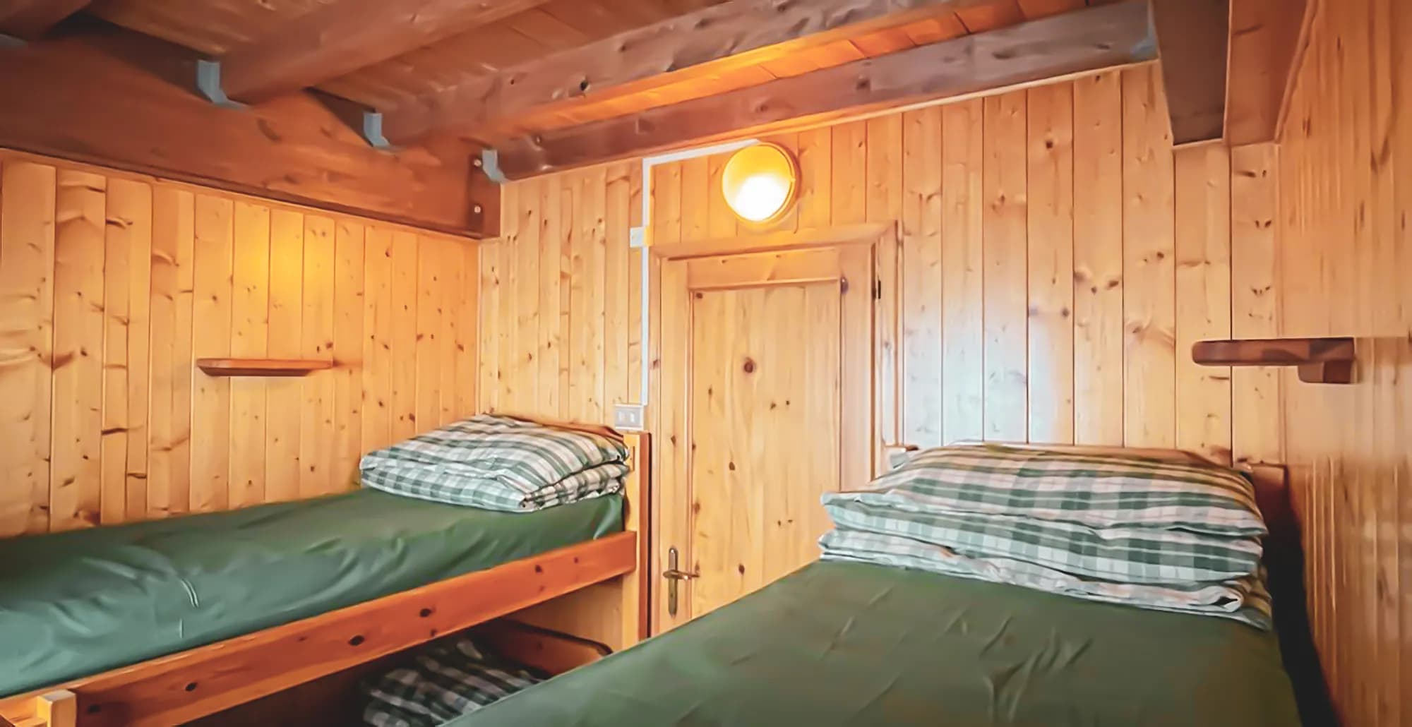 A wooden bedroom with two bunk beds. The walls and ceiling are covered in wood panels, creating a warm, rustic atmosphere. The beds are covered with a green sheet, and the pillows are covered with a white sheet.