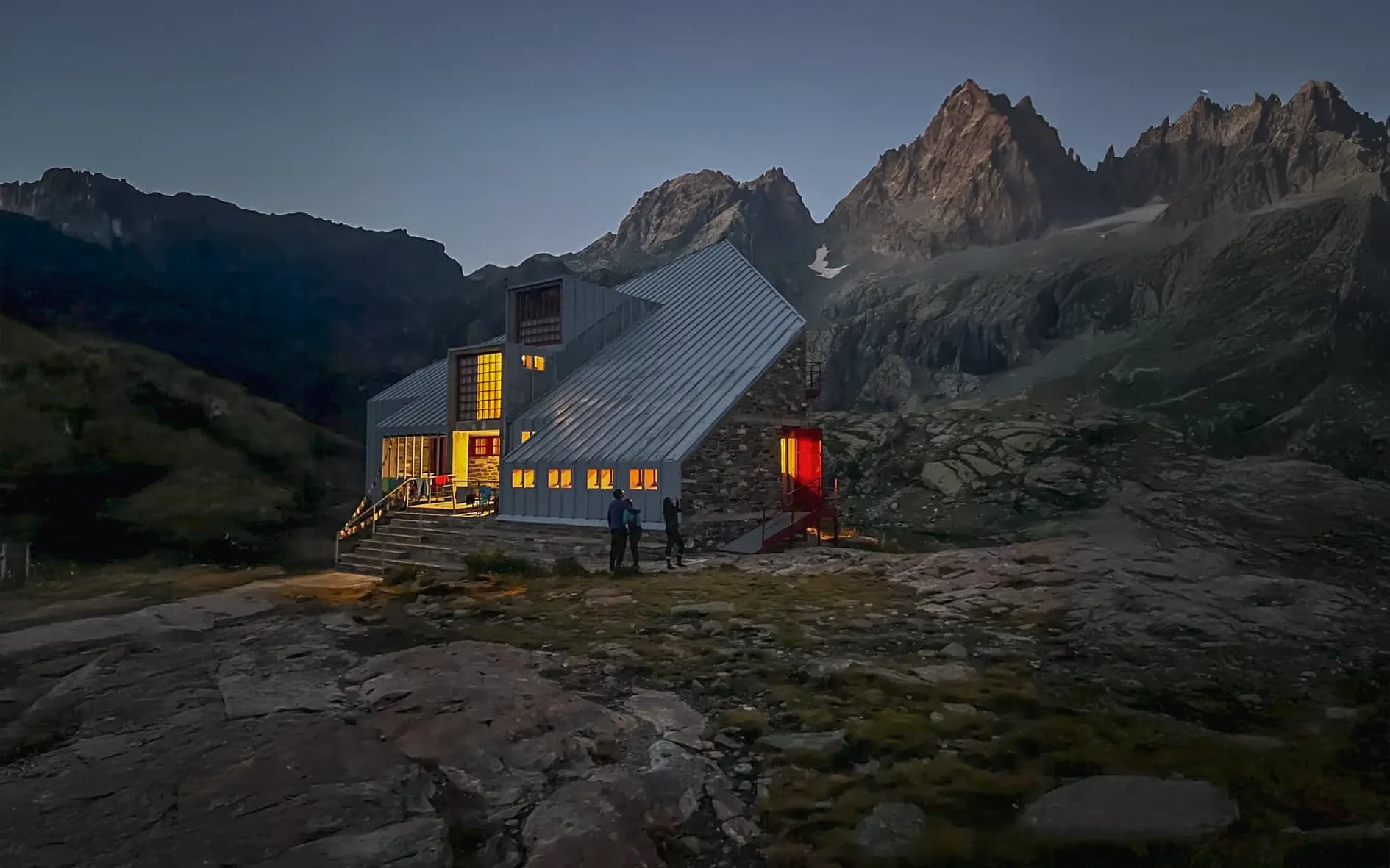 A modern Mountain hut lit up at dusk, surrounded by majestic mountains. A wilderness adventure.