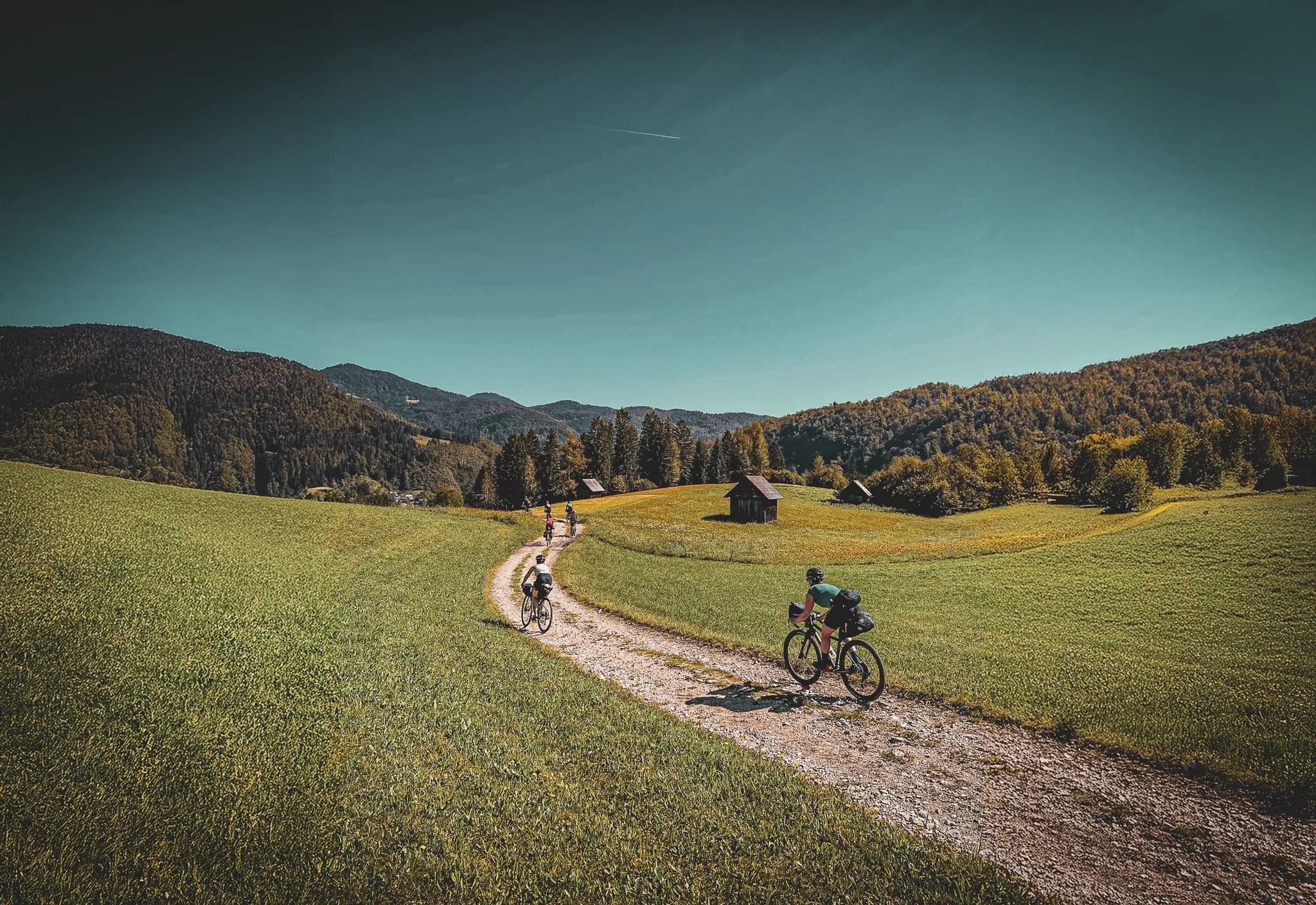 A winding road cuts through a green landscape, with cyclists pedalling along a stony path. In the background, green hills and colourful trees rise up under a clear sky.