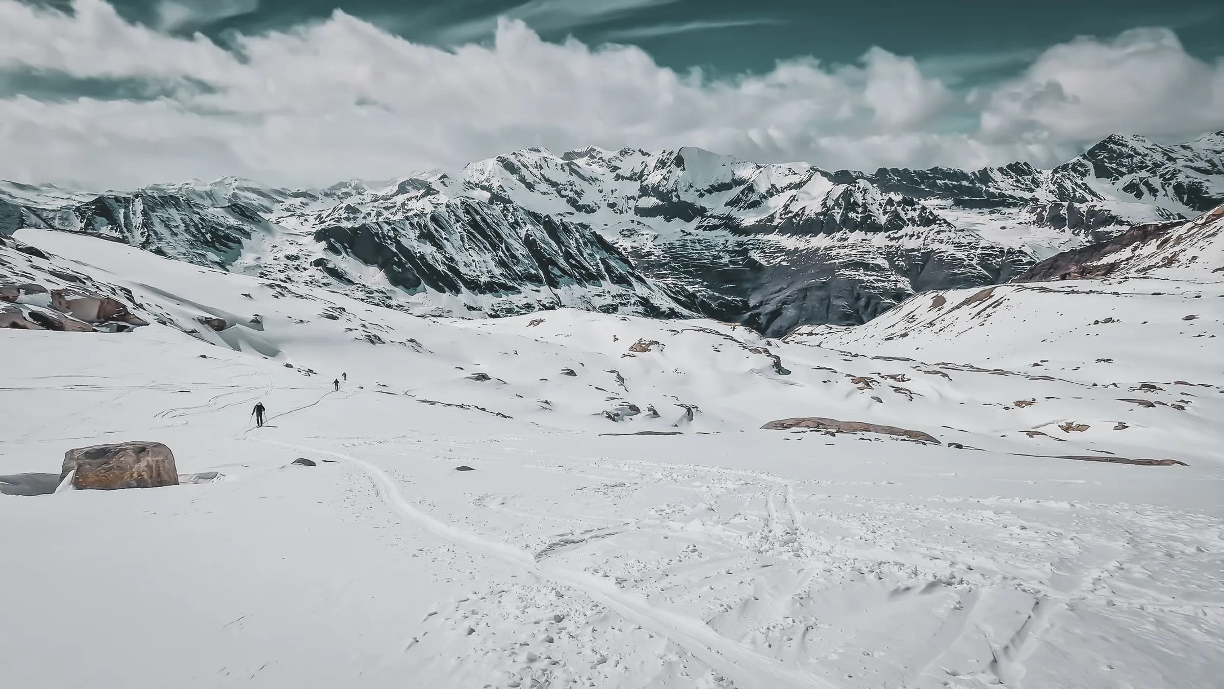 A majestic snowy landscape with imposing mountains in the background. The ground is covered with a thick layer of snow, creating a pure and peaceful atmosphere. Several alpine silhouettes can be seen.