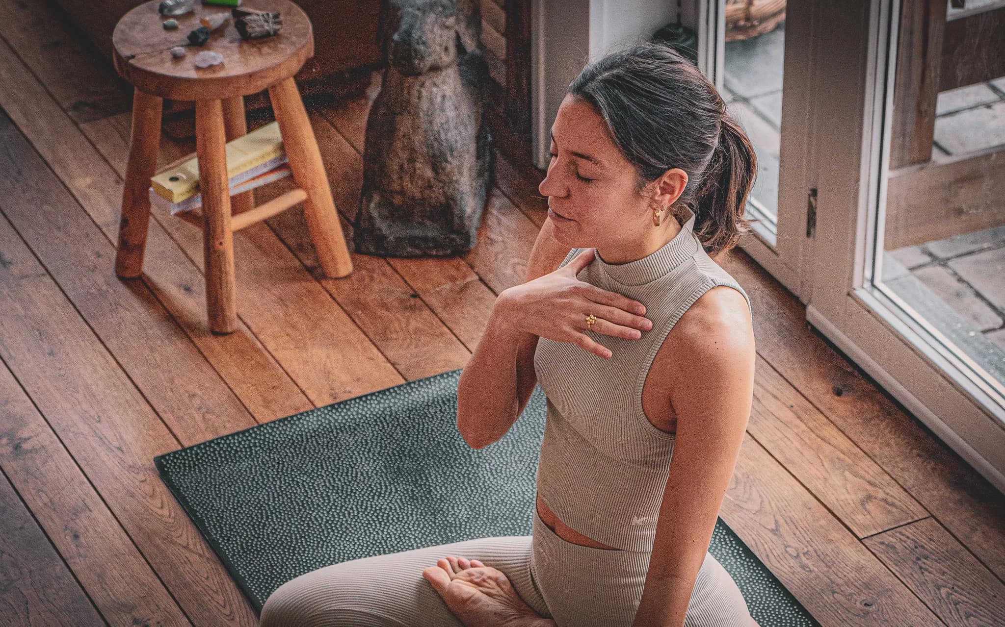 A young woman practises meditation in a warm interior, sitting on a yoga mat. She is wearing a tight beige sports outfit, with her hair pulled back. Her posture is calm and concentrated, with one hand on her back.