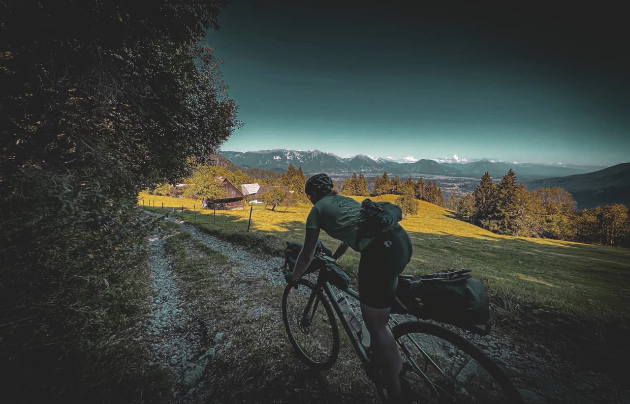A cyclist climbs a gravel road in a mountainous region, surrounded by green meadows and trees. Wooden chalets can be seen in the background, giving the scene a rural feel.