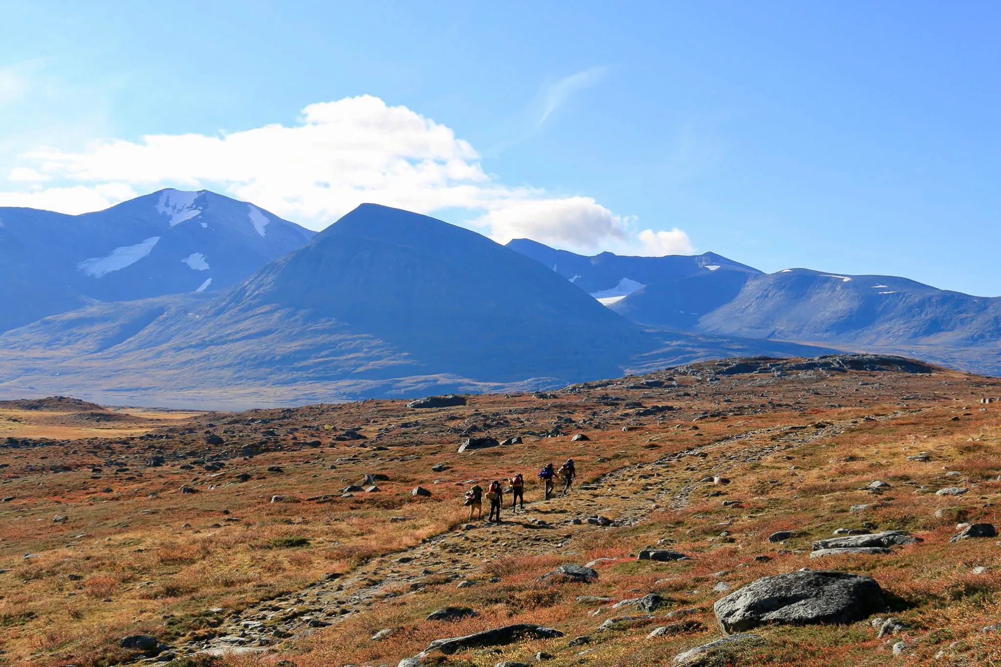 A mountainous landscape in a wilderness area with hills and mountains in the background, partly covered in snow. The sky is a light blue, dotted with white clouds. In the foreground, a group of