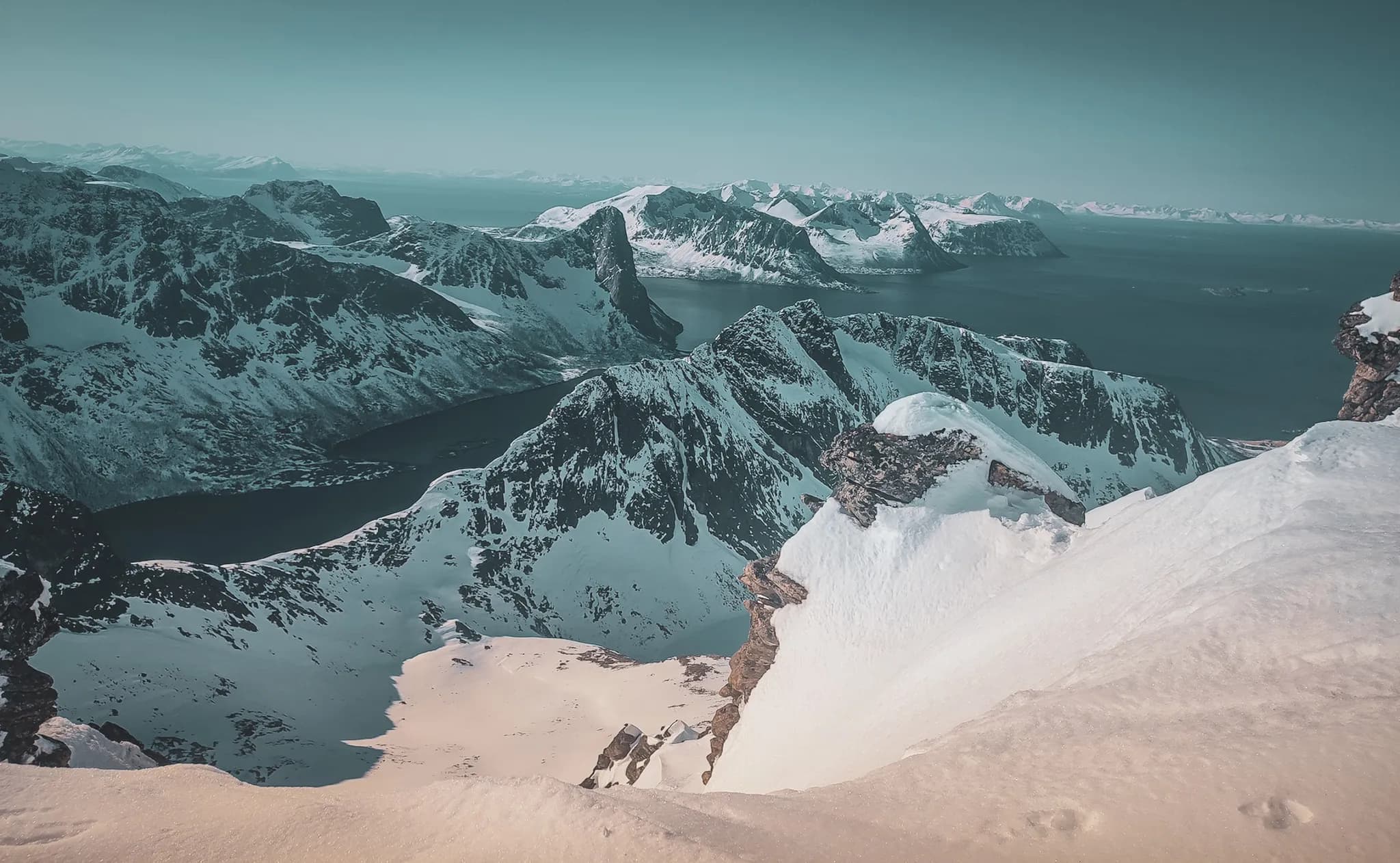 Een spectaculair panorama van majestueuze besneeuwde bergen die uittorenen boven een sprankelende fjord. De gevarieerde vormen van de toppen rijzen trots de lucht in in witte en grijze tinten, terwijl