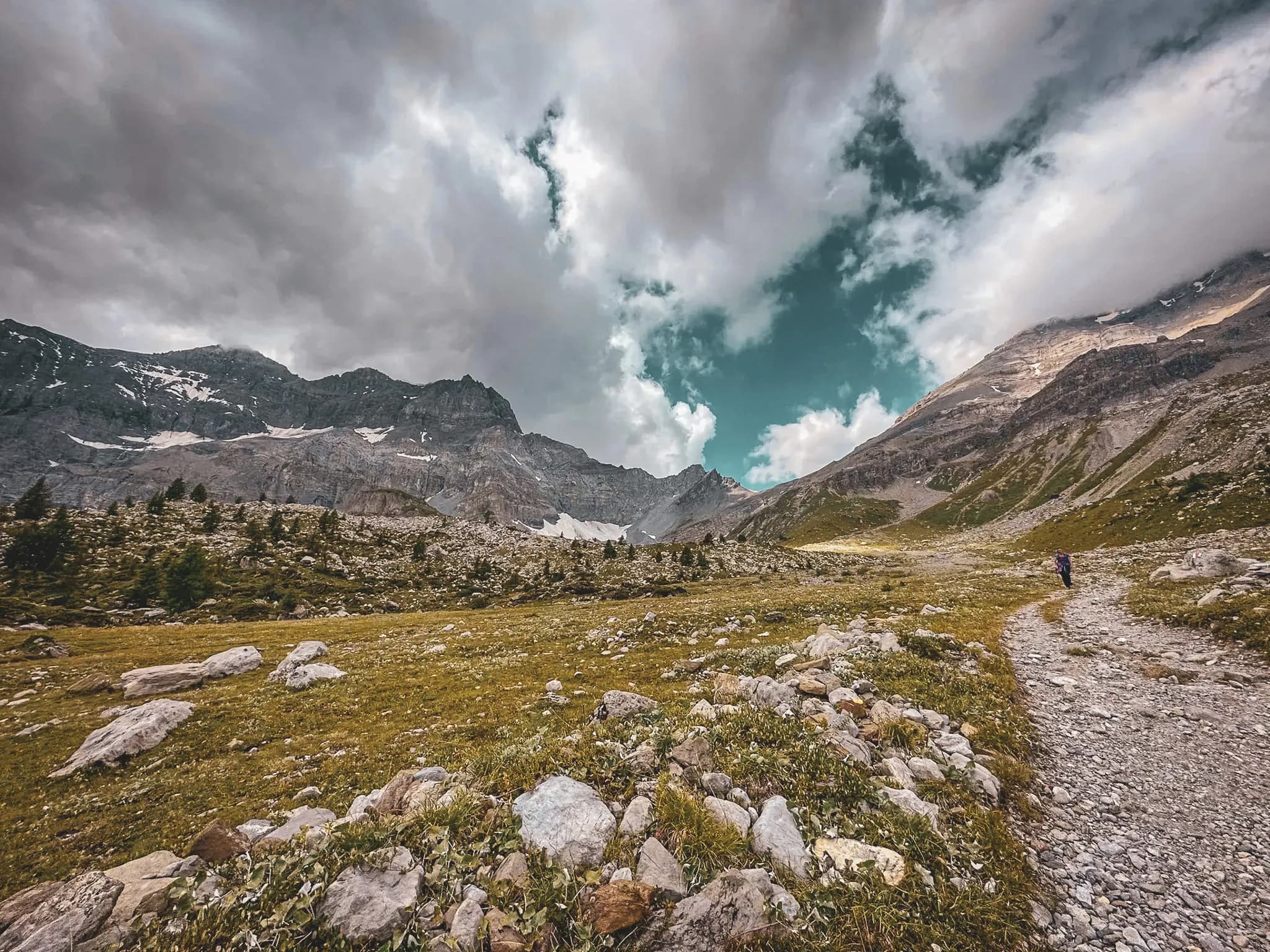 A vast, serene mountain landscape, characterised by steep peaks partly covered in snow. The sky is cloudy, with flashes of blue between the clouds. A scent