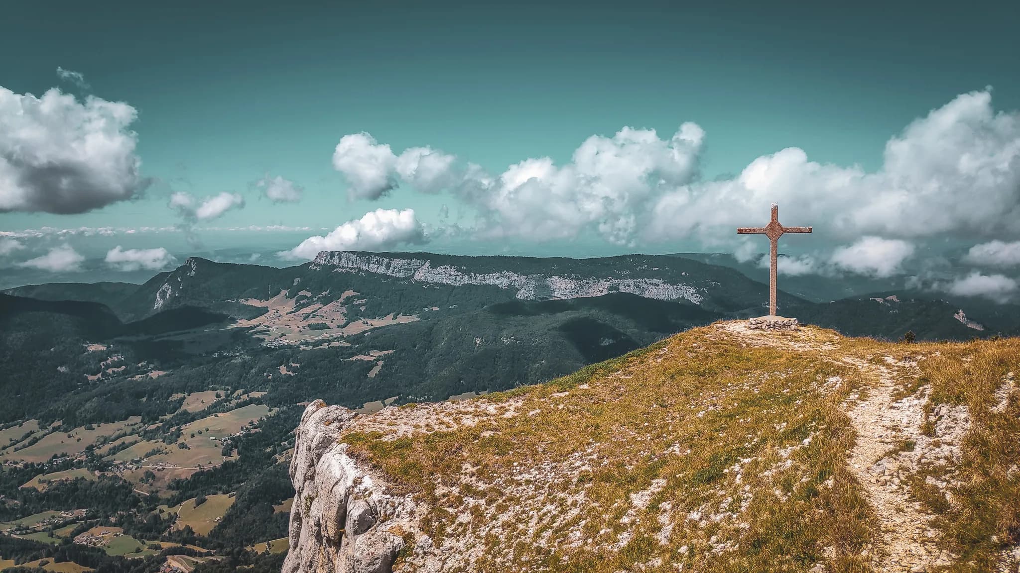 Un sommet de montagne avec une croix en bois, entouré de verdure et surplombant un paysage vallonné. En arrière-plan, des collines et des falaises se dessinent sous un ciel bleu parsemé de nuages