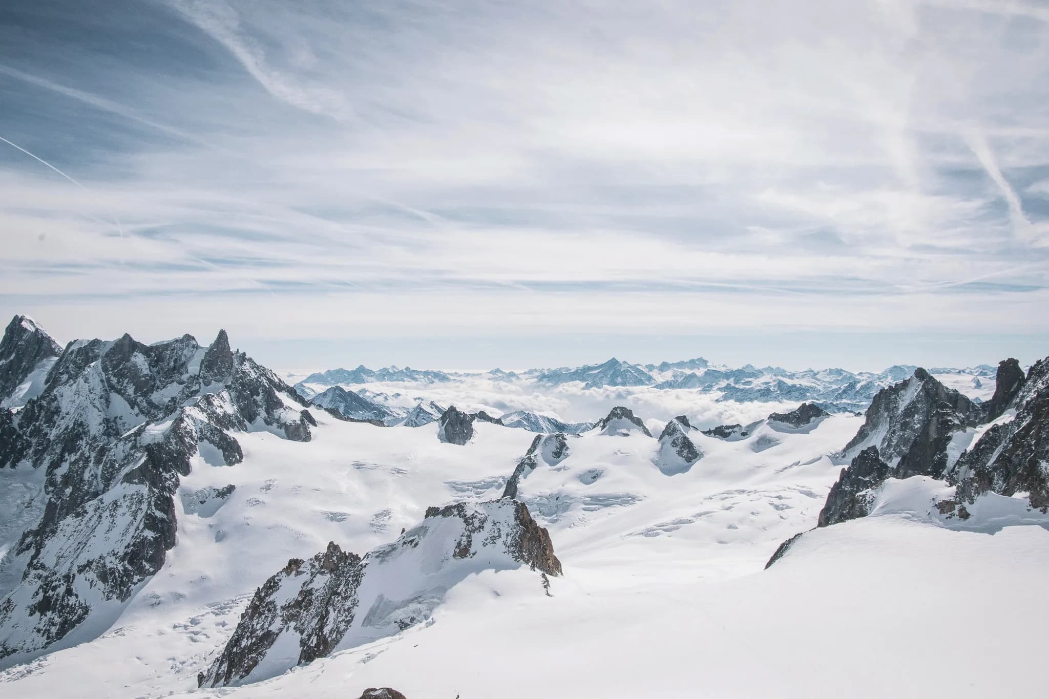 Un paysage de montagnes enneigées, avec des sommets rocheux se dressant dans un vaste décor blanc. La neige recouvre le sol tout autour, créant une surface lisse et uniforme, tandis que des formations rocheuses émergent ç