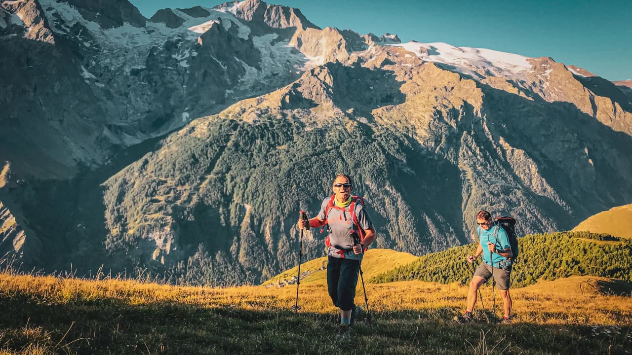 Deux randonneurs souriants progressent sur un sentier alpin, entourés de majestueuses montagnes.