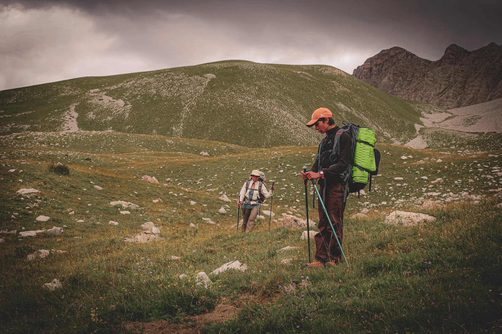 A landscape of green mountains with two hikers on a rocky terrain. One of the hikers, wearing a dark t-shirt and carrying a green rucksack, appears to be concentrating on his task.