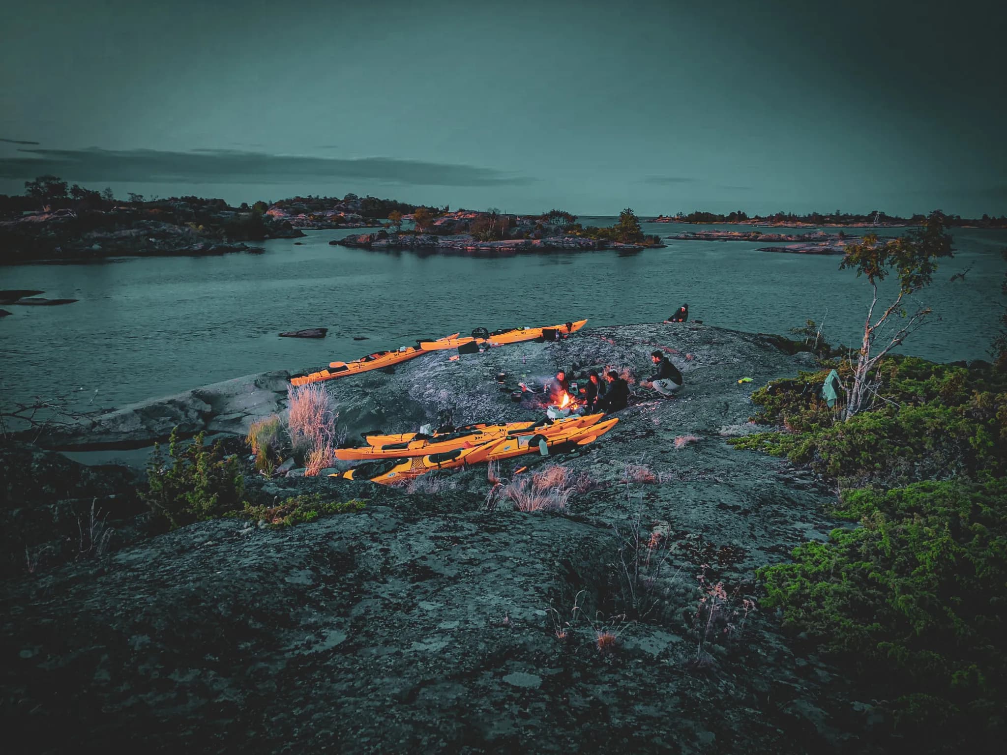 A group of people are gathered around a campfire on a rocky surface near a stretch of water. Several bright yellow kayaks are moored on the shore, adding a colourful touch to the scene.