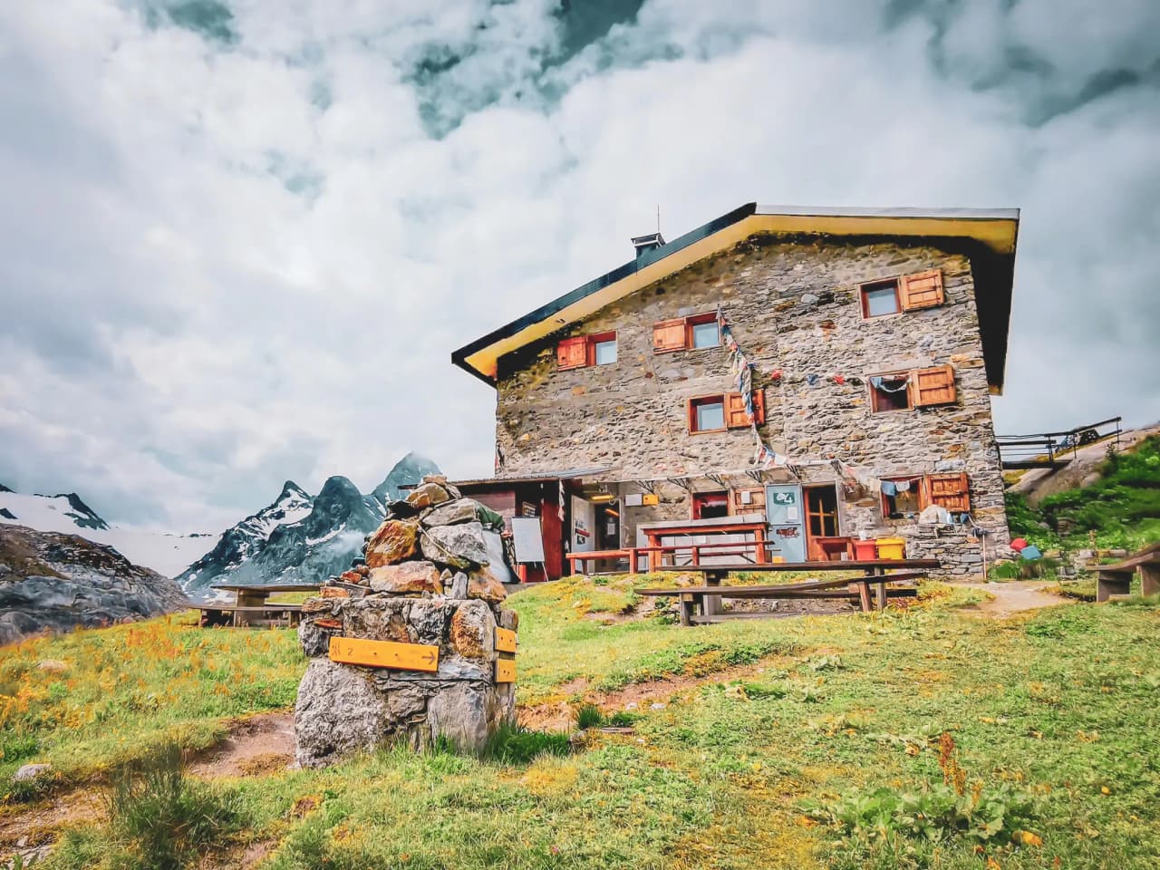 Cabane en pierre perchée dans les Alpes, face aux montagnes, entourée d'herbe et de fleurs.