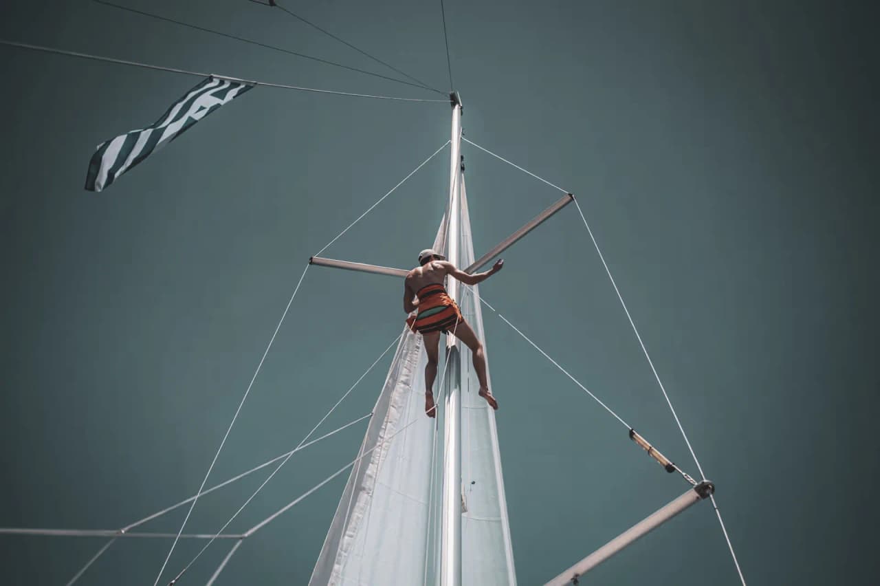 Un grimpeur monté au mât d'un voilier sous un ciel bleu, prêt pour l'aventure méditerranéenne.
