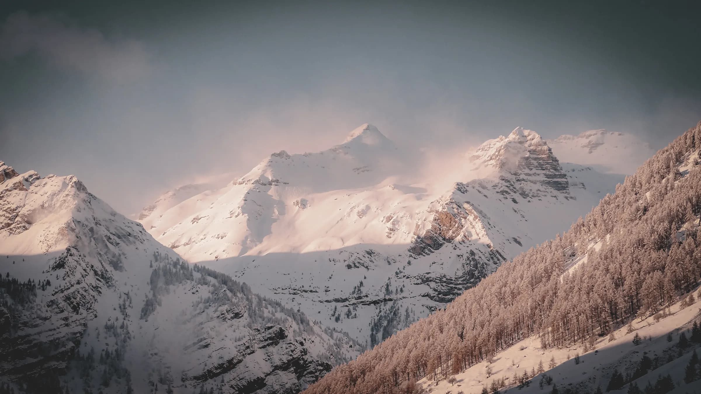 A spectacular mountain landscape, with snow-capped peaks towering above. The mountains, covered in a thin layer of snow, are illuminated by a soft light, creating a contrast between the steep slopes and the peaks.