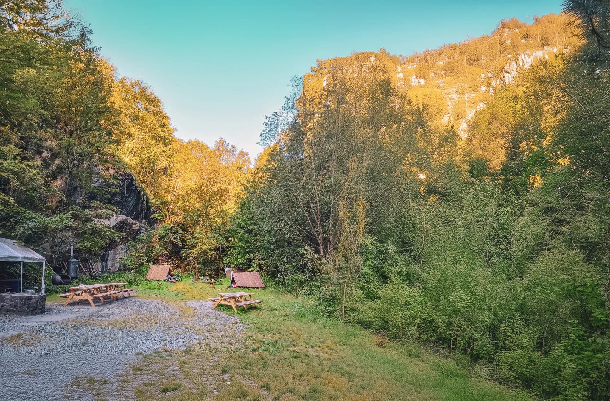 A space in the middle of nature, with wooden tables laid out on a gravel floor. On the left, a shelter with a canvas roof and a fireplace, while smaller structures, probably tents, can be seen behind.