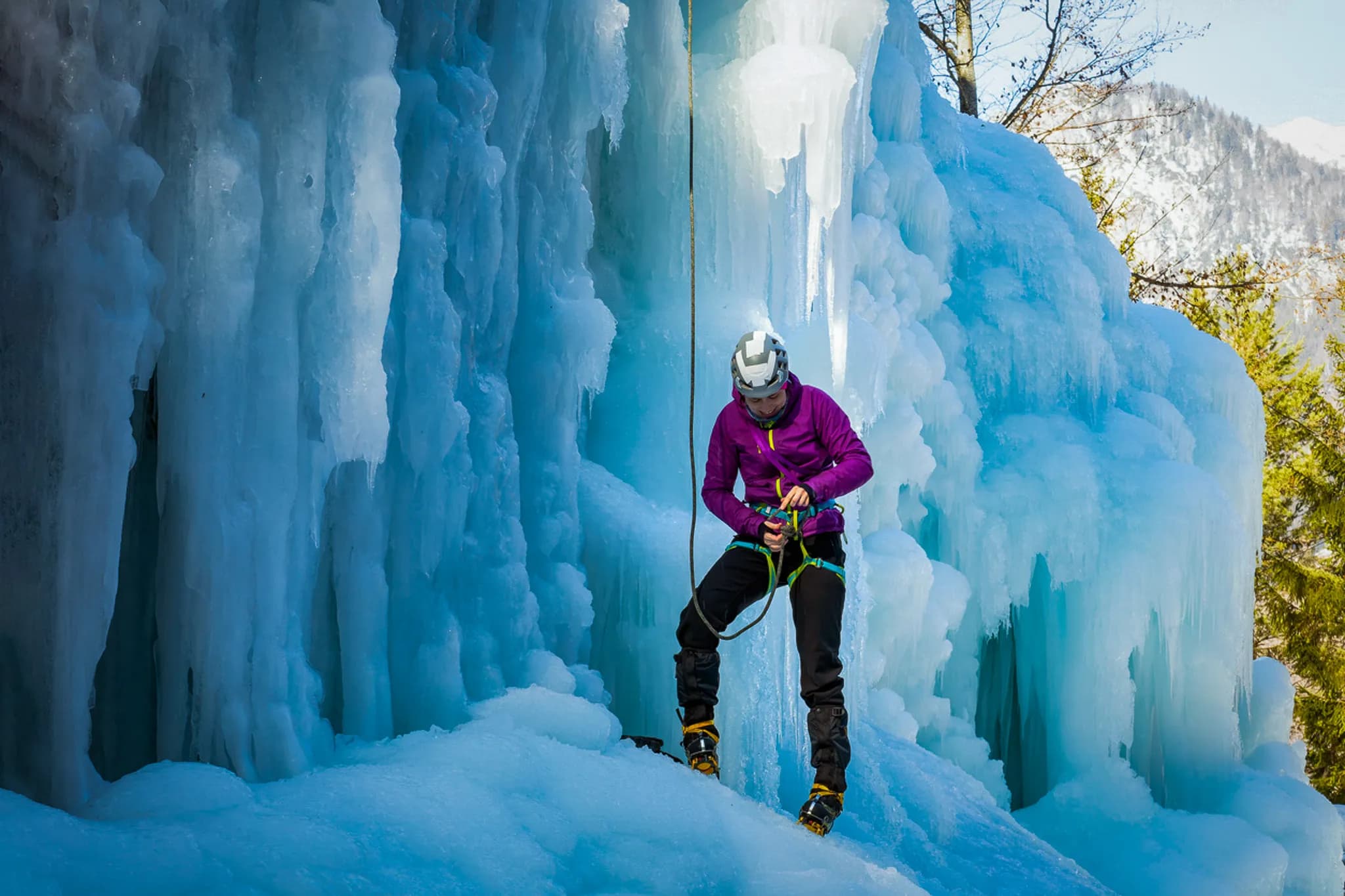Un grimpeur en plein effort escalade une paroi de glace, entouré de formations de glace bleuâtres et scintillantes. Il porte un casque de protection et une veste violette, se préparant à utiliser son équipement avec des