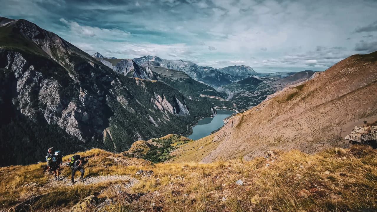 Un aperçu époustouflant des montagnes avec randonneurs, verdure et lac scintillant sous le ciel bleu.