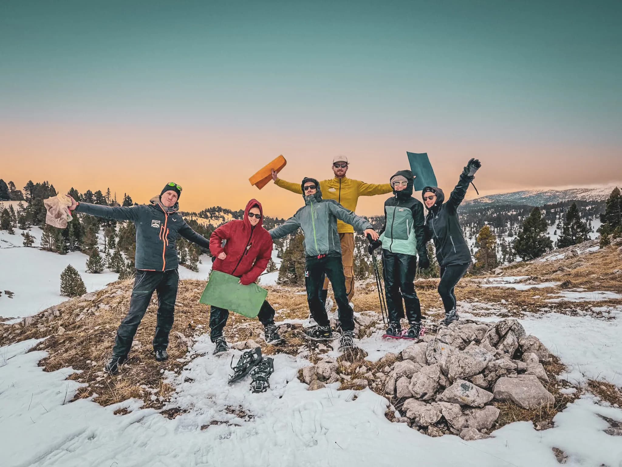 A group of six people stand on a snow-covered hilltop, smiling and celebrating. They are dressed in winter-appropriate outdoor clothing, ranging from colourful jackets to accessories such as a snowman's jacket.