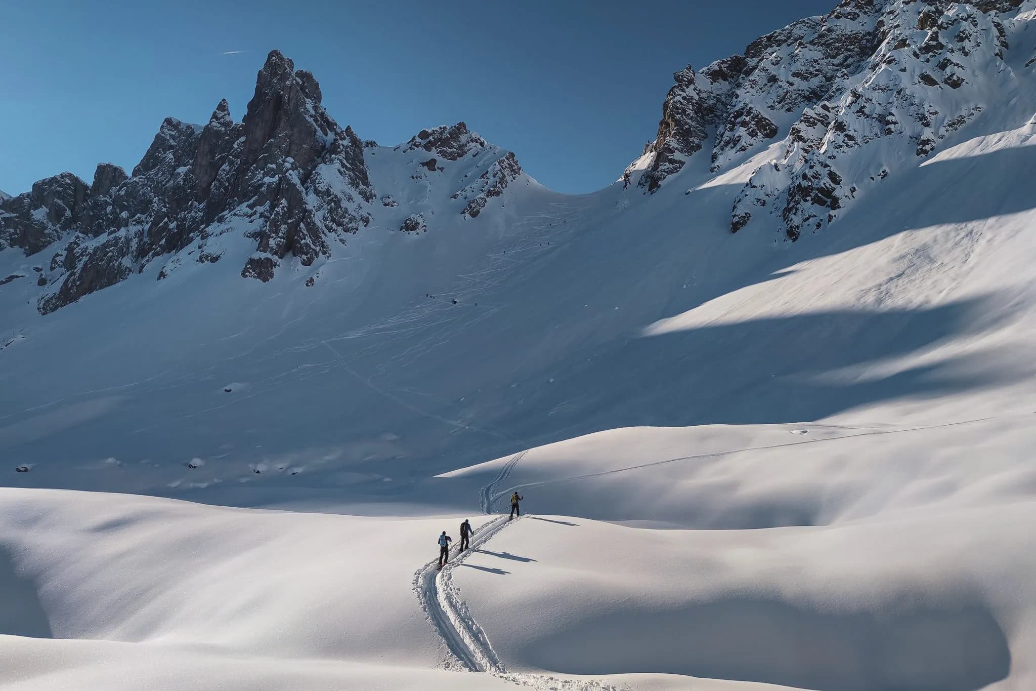 Un paysage alpine recouvert de neige, avec des sommets montagneux escarpés en arrière-plan. Sur la surface immaculée, plusieurs randonneurs avancent en suivant des traces laissées dans la neige. Le ciel est clair