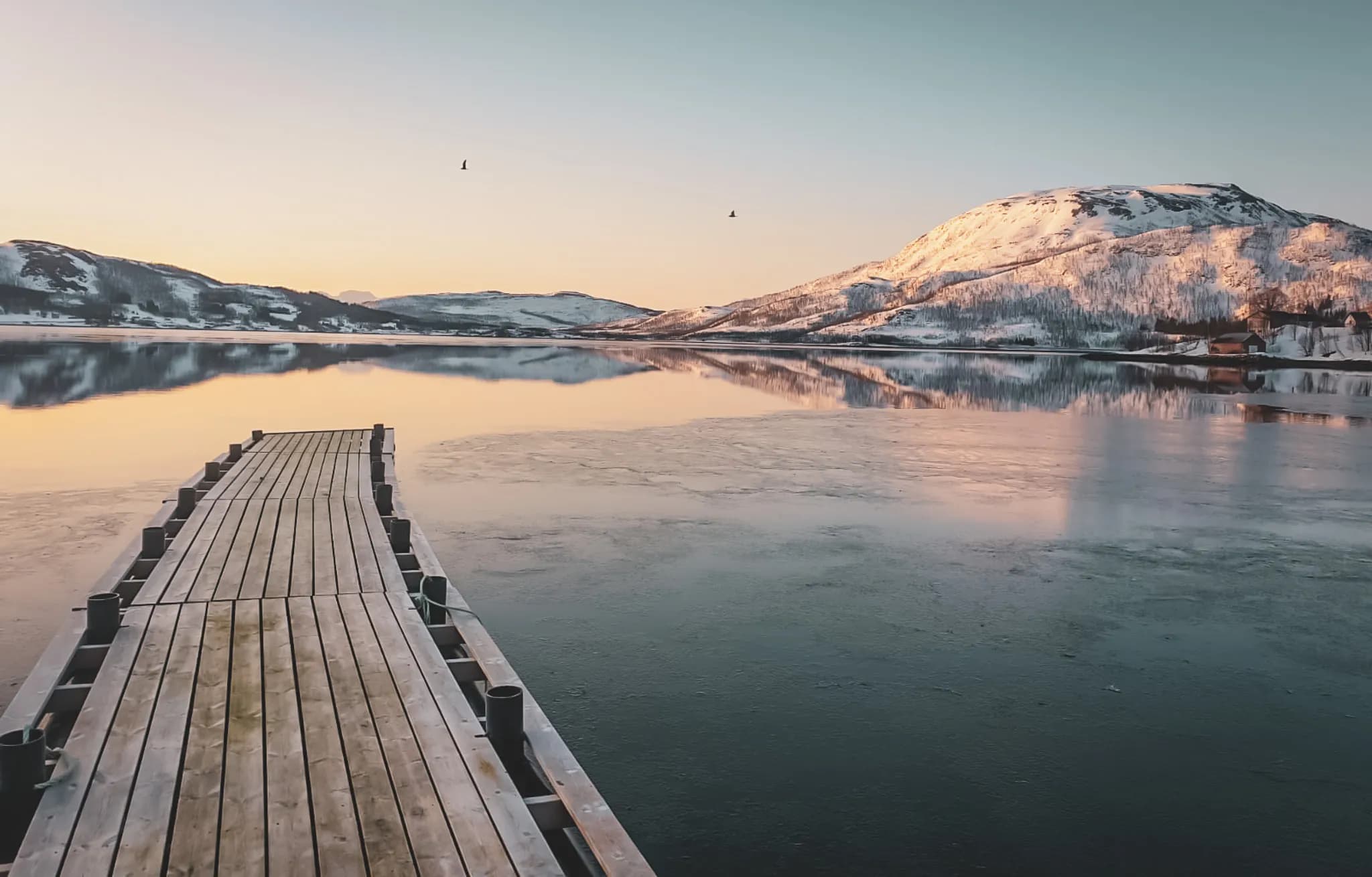 Een houten steiger strekt zich uit over een kalm meer, omringd door besneeuwde heuvels. De lucht is getint met pasteltinten die doen denken aan dageraad of schemering. Reflecties van bergen en