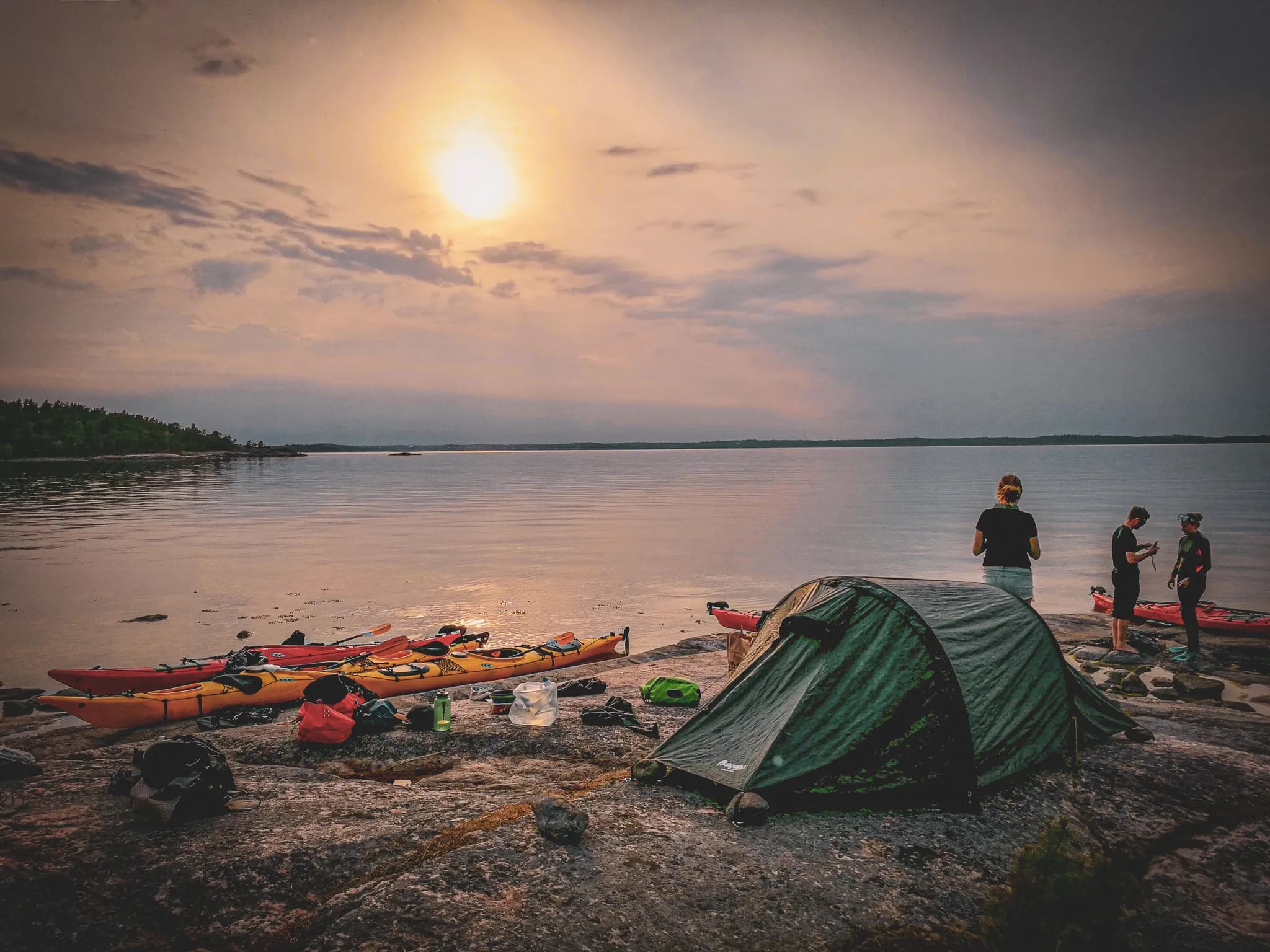 A tranquil coastal landscape at dusk, with a calm lake reflecting the light of a setting sun. A small group of people stand near an encampment, consisting of a green tent set on rocks.