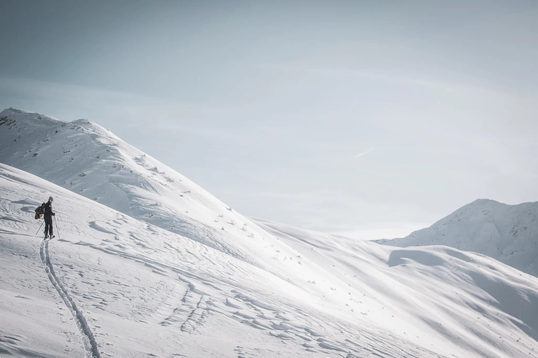 Un skieur trek sur une vaste étendue recouverte de neige, ascendant une pente douce d'une montagne. La neige est immaculée, parsemée de traces laissées par ses skis. En arrière-plan, des sommets montagneux