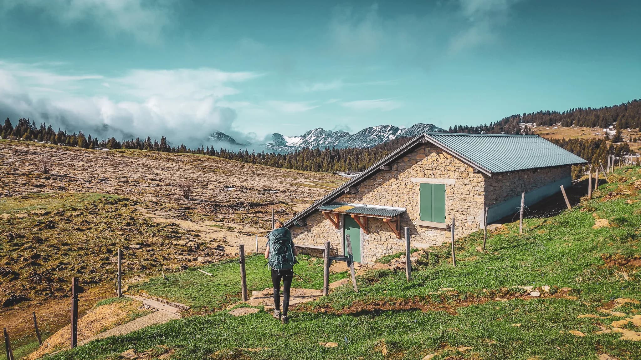 Un chalet en pierre se trouve au milieu d'un paysage montagnard, avec une vue dégagée sur des sommets enneigés en arrière-plan. Au premier plan, un sentier verdoyant mène à l'entrée du chalet, entouré de