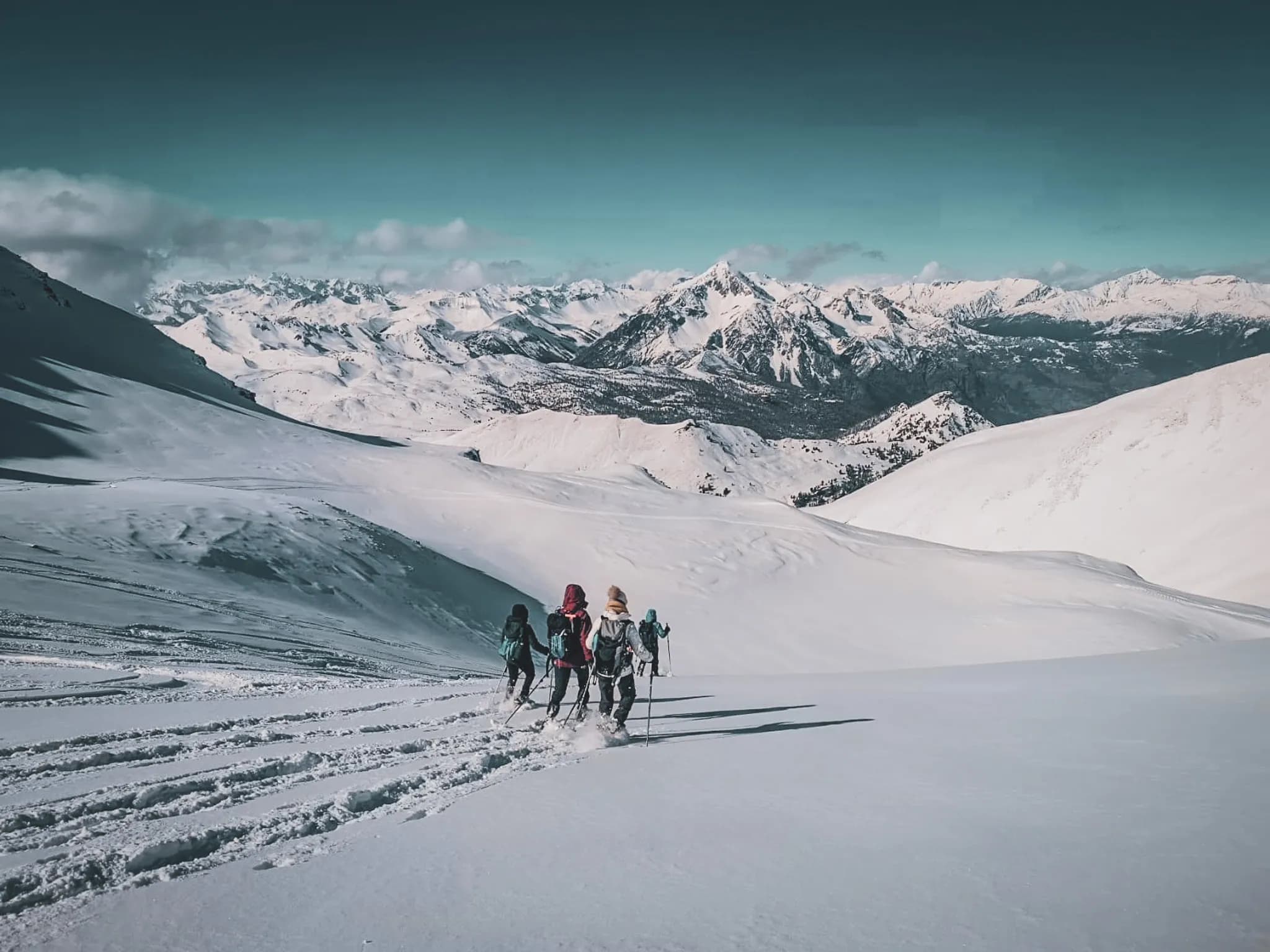 A snow-covered mountain landscape, with several hikers making their way along a gentle slope. The majestic mountains in the background are dotted with snow and bathed in a slightly cloudy sky.