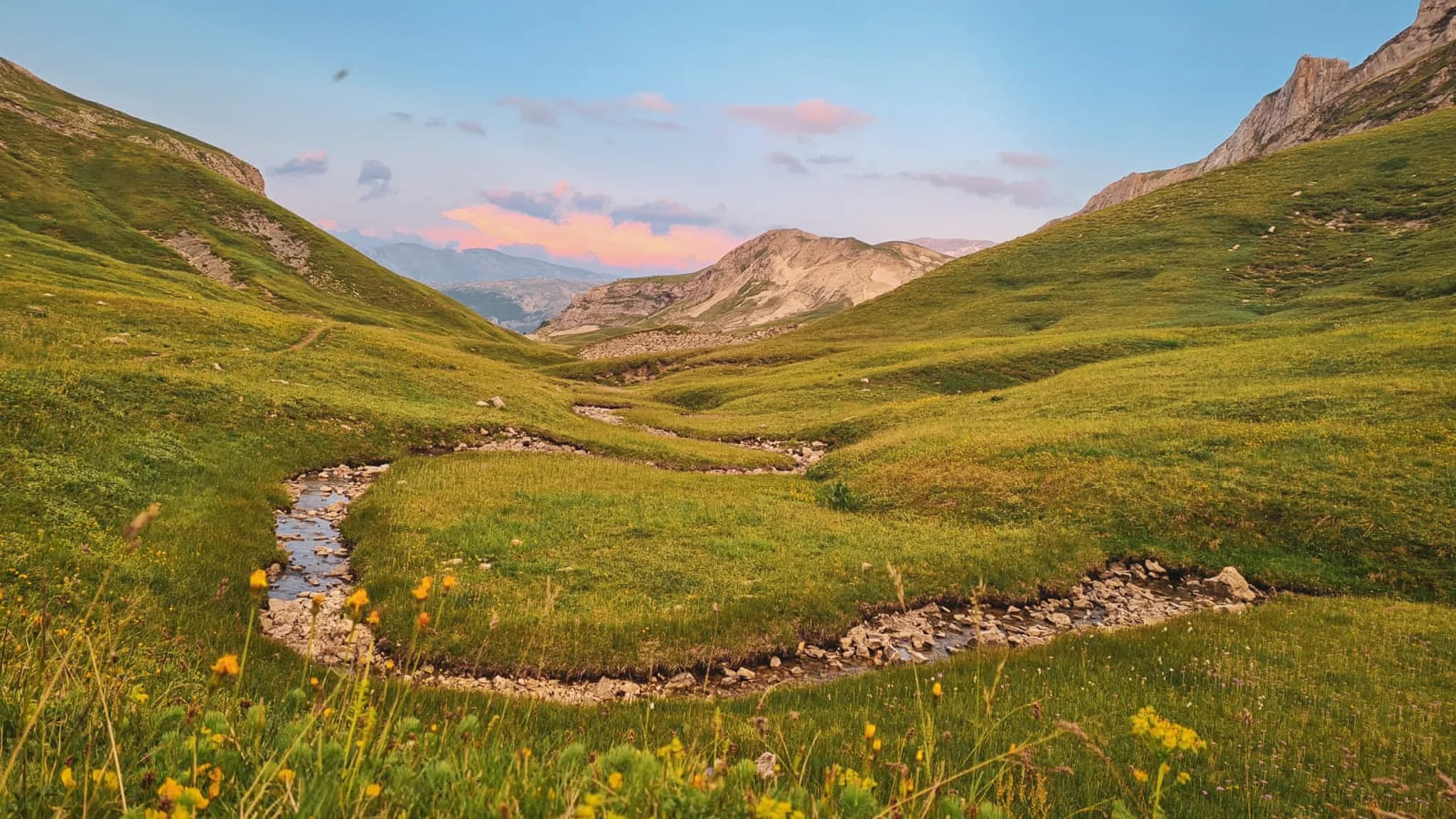 Vast green meadows in the heart of the Alps, crossed by a sparkling stream.