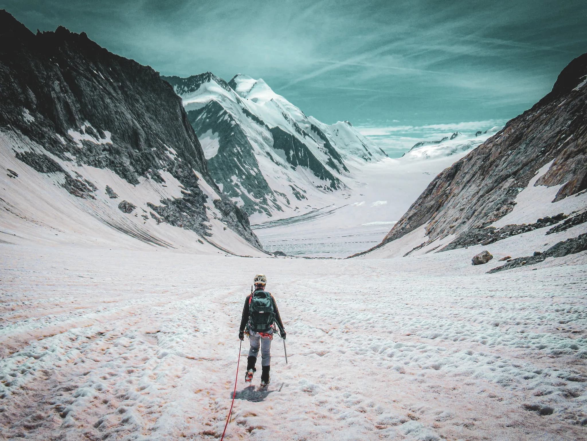 An equipped mountaineer heads for a vast glacial landscape, with majestic mountains in the background. The ground is covered in snow and ice, and the sky has turquoise hues.