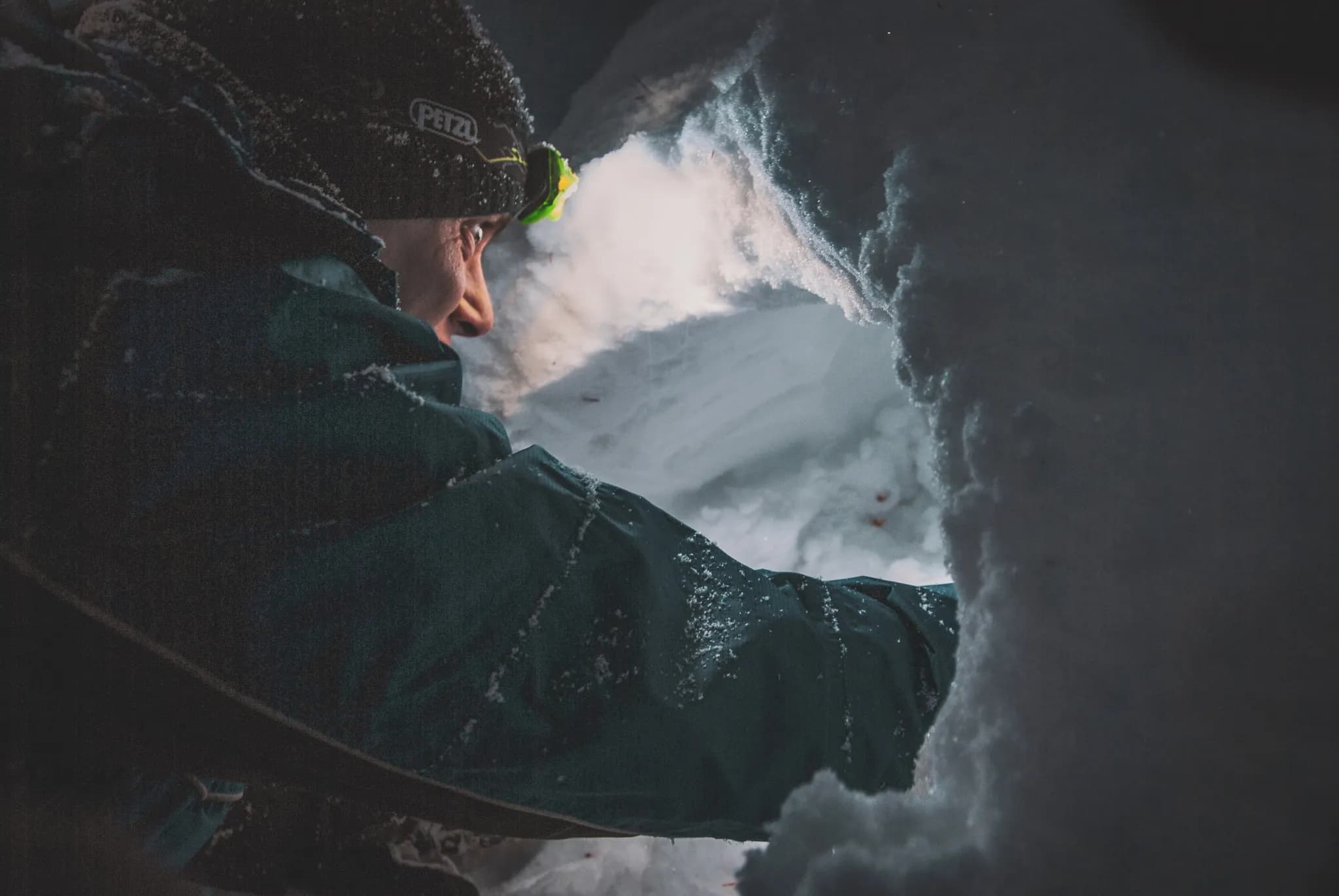 A man dressed in a winter jacket and cap is leaning over a hole in the snow. He uses a headlight to illuminate the inside of the snow-covered shelter, highlighting the texture and texture of the snow.