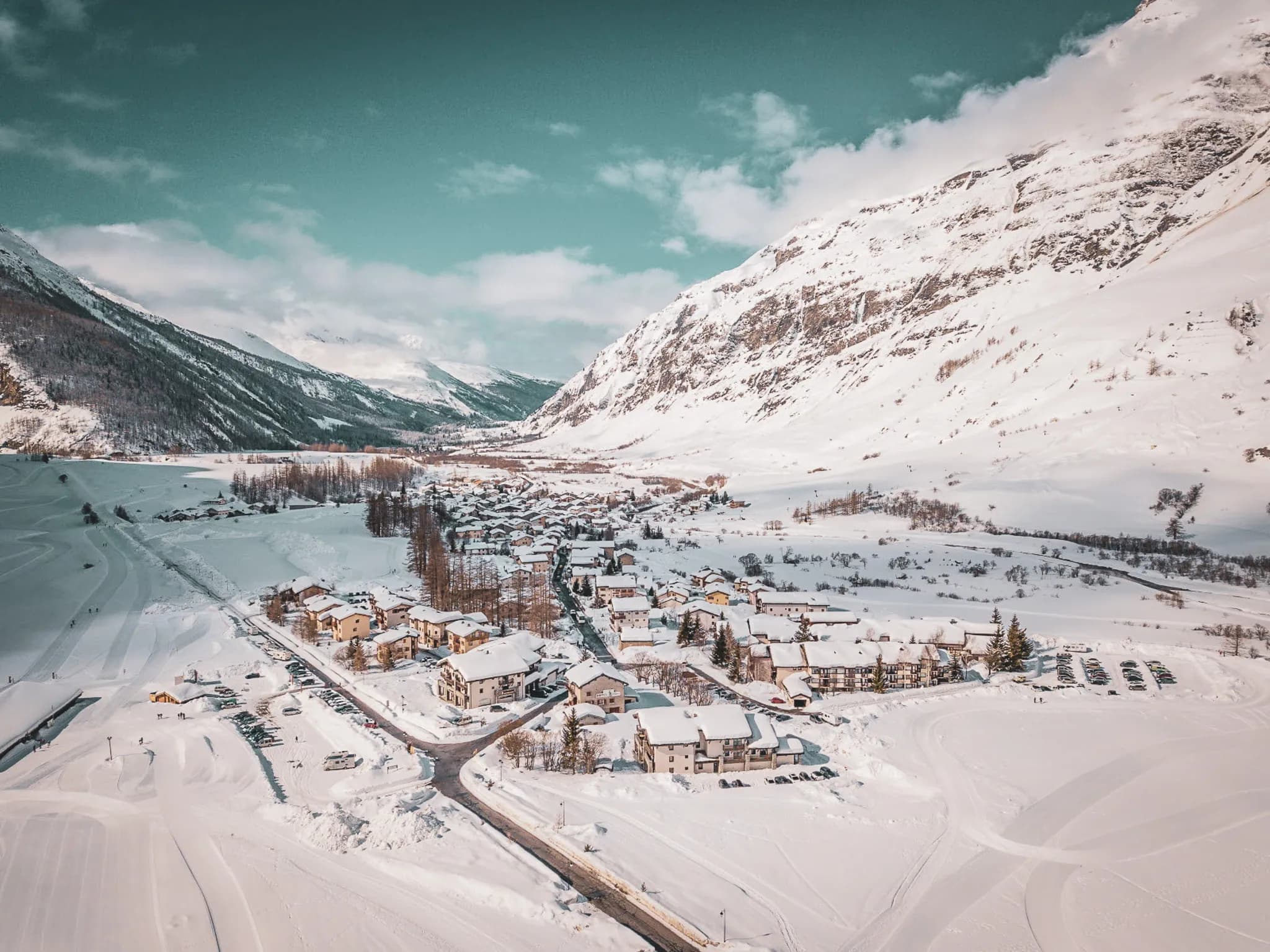 Un village enneigé entouré de montagnes majestueuses. Des bâtiments colorés et traditionnels sont dispersés dans la vallée, avec des toits recouverts de neige. De larges étendues de neige immaculée s'ét