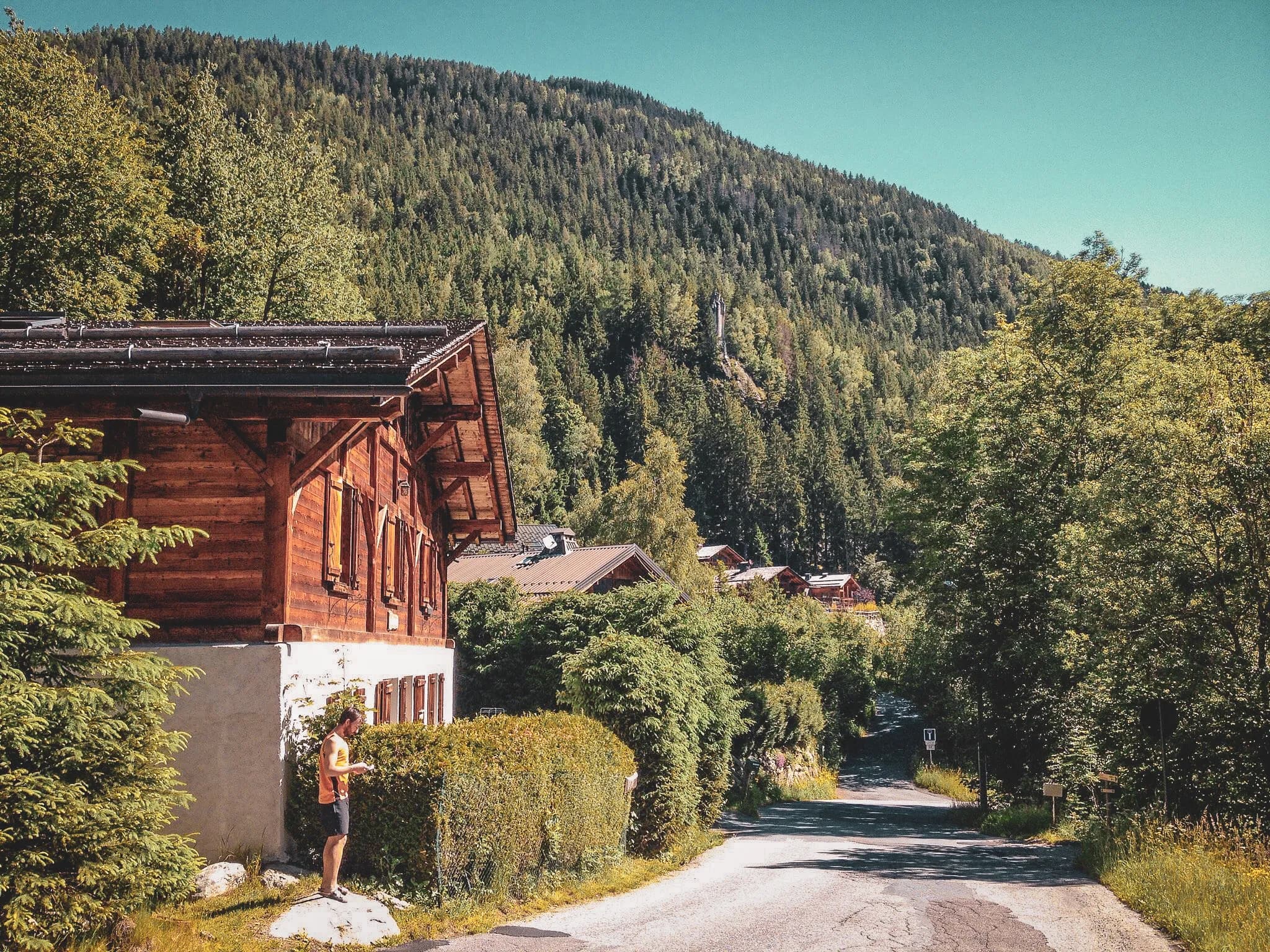 A wooden cottage on the side of a winding road, surrounded by lush greenery. A man stands by the chalet, watching his phone. In the background, majestic mountains with dense forests of