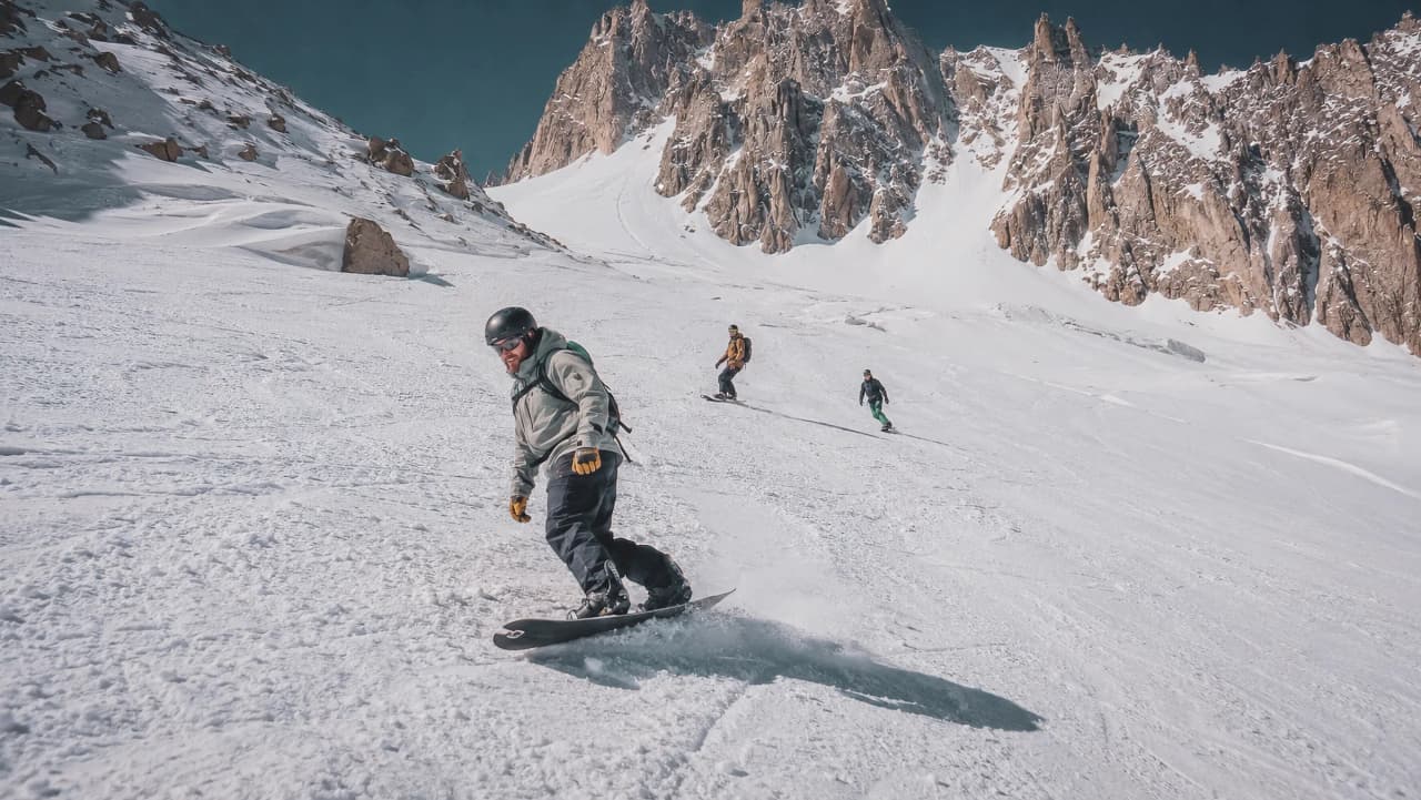Un snowboardeur descend des pentes enneigées avec des sommets majestueux en arrière-plan.
