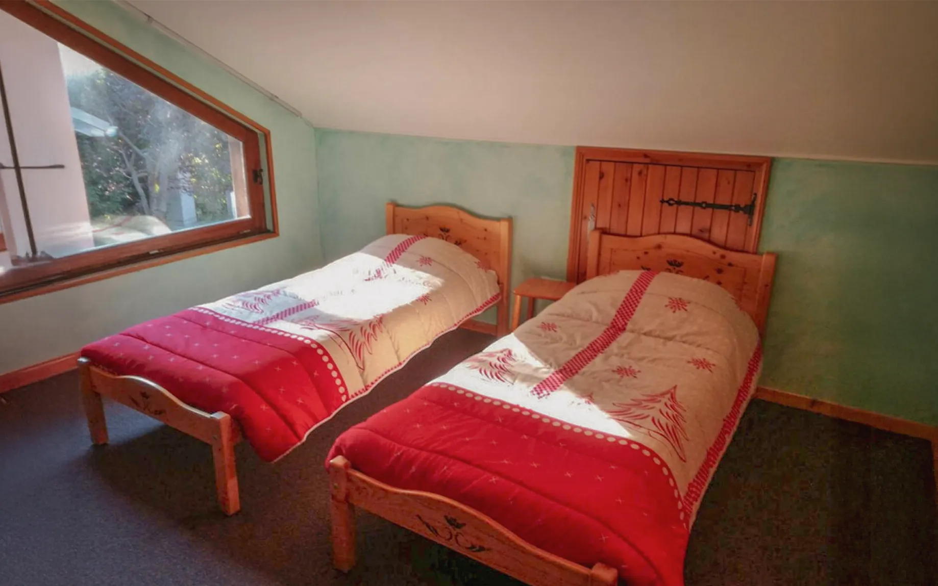 A bedroom in a chalet, with two single wooden beds, each covered with a duvet with red and white motifs. The room is bright thanks to a large window with a view of the outside. The