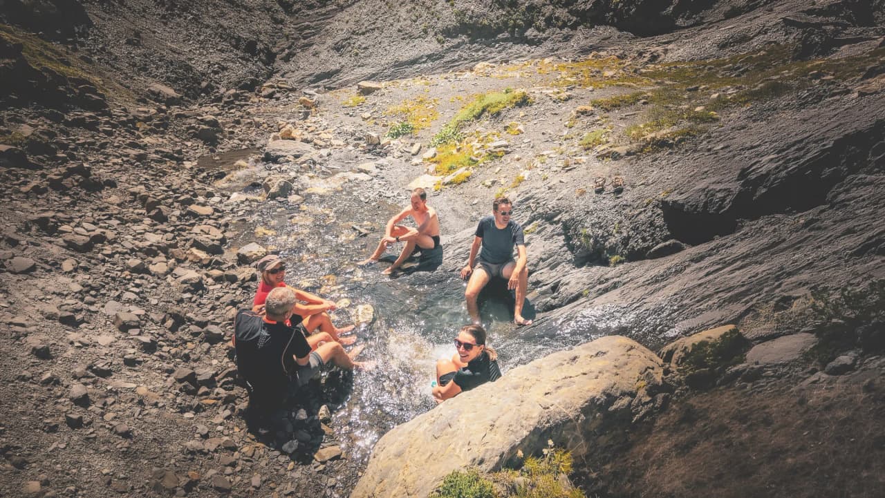 Groupe d'amis se relaxant dans un ruisseau en montagne, entouré de pierres et de verdure.