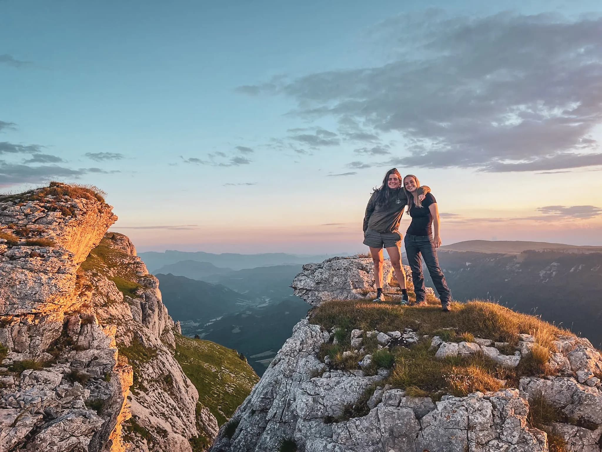 Two smiling hikers on top of a rock, with a spectacular mountain panorama at sunset.