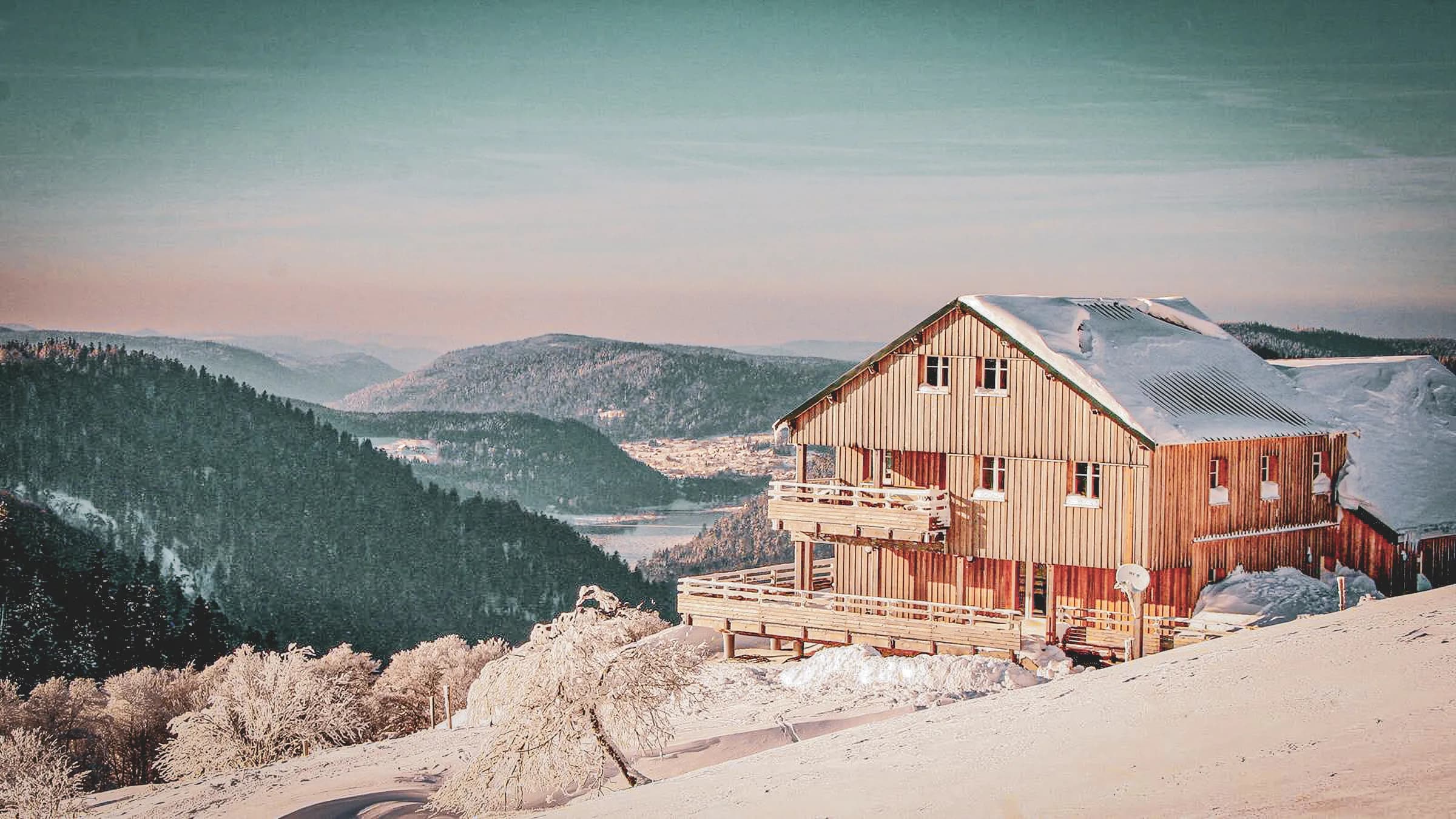 Een houten chalet staat in een besneeuwd landschap, met bergen op de achtergrond. Het twee verdiepingen tellende gebouw is gedeeltelijk bedekt met sneeuw en heeft een ruim balkon. De omringende natuur is