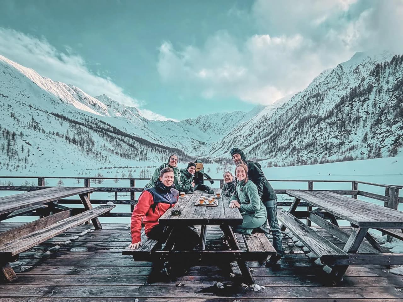 Groupe d'amis souriants autour d'une table en bois, avec les montagnes enneigées en arrière-plan.