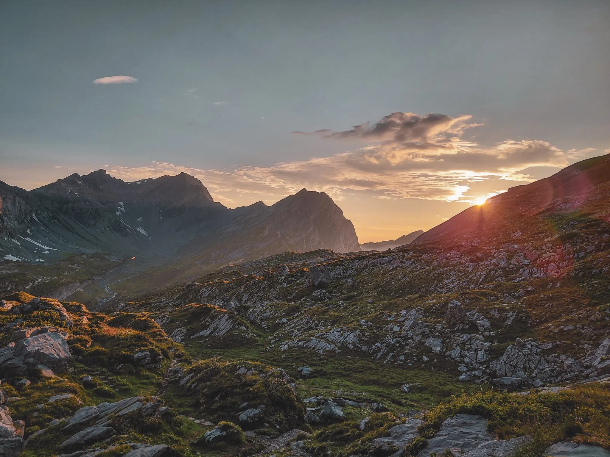 A spectacular mountain landscape, with undulating rock formations covered in dark gravel. In the background, majestic peaks loom, bathed in soft light. To the right, a lake