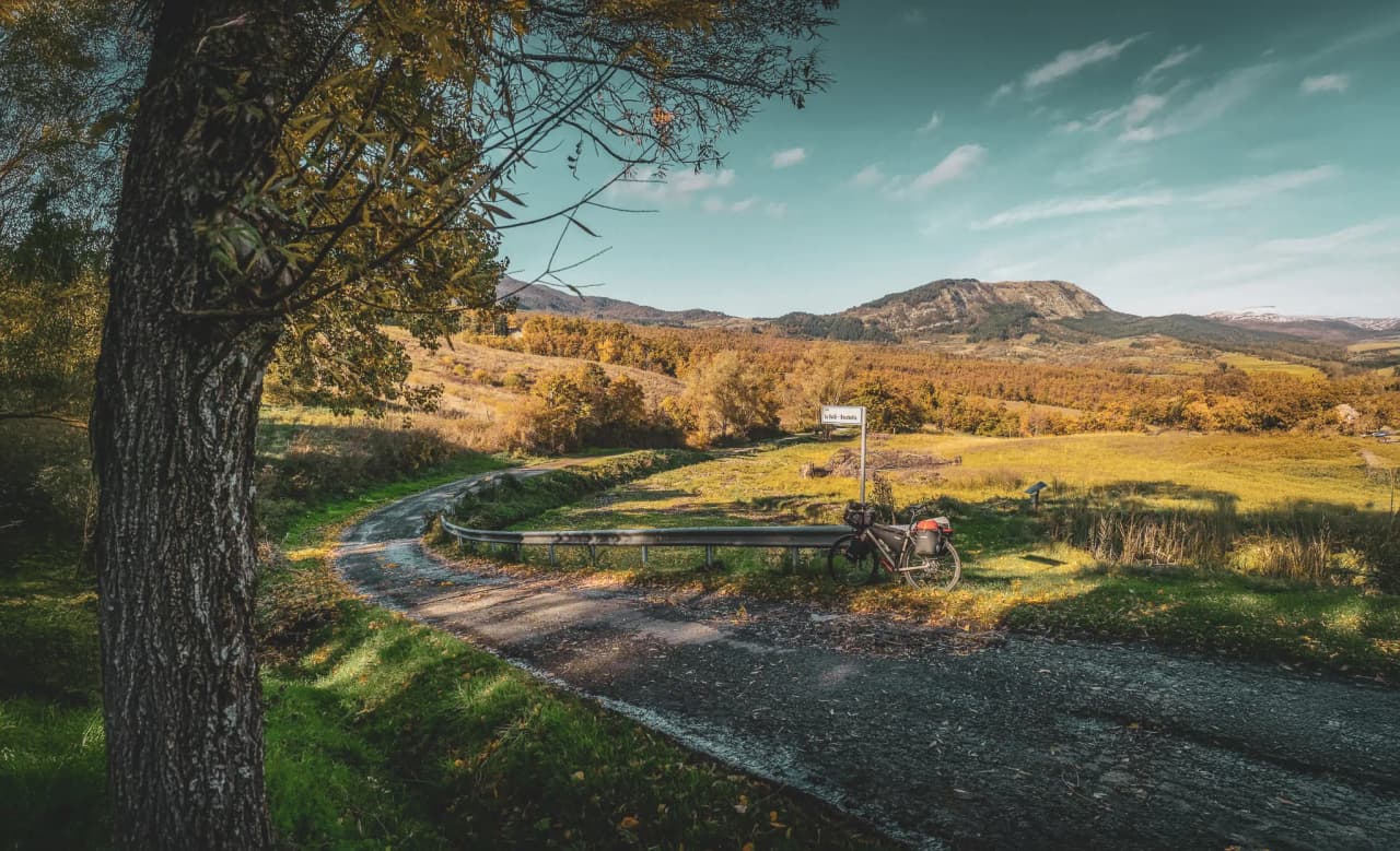 A winding road leads through the Casentinesi area, surrounded by hills and mountains in the background. The scenery is reminiscent of autumn, with golden-leafed trees.