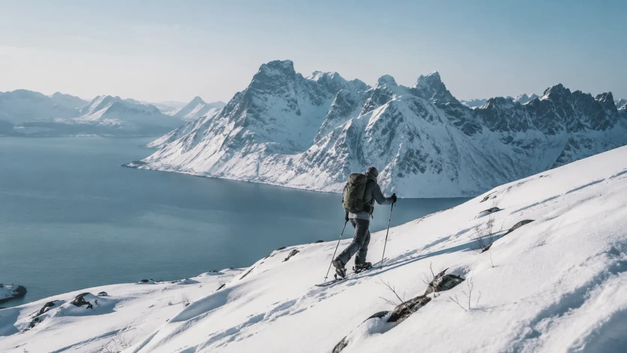 Snowshoe hiker on a snow-covered fjord landscape in Senja, Norway, under a serene sky.