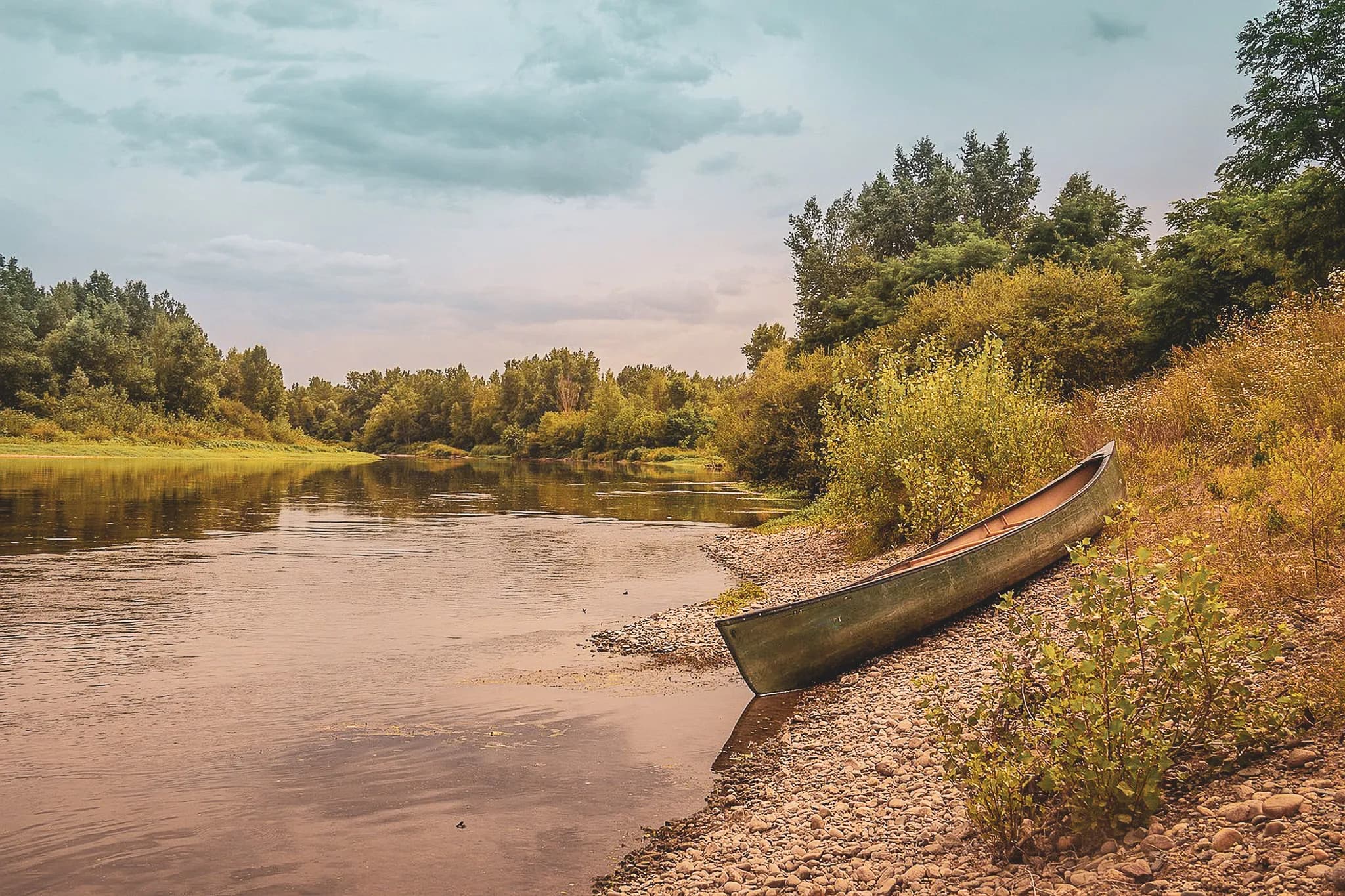 A calm natural landscape with a river lined with green trees. On the shore, a wooden canoe is moored on a pebble beach. The surface of the water is gently undulating, reflecting the sun's rays.