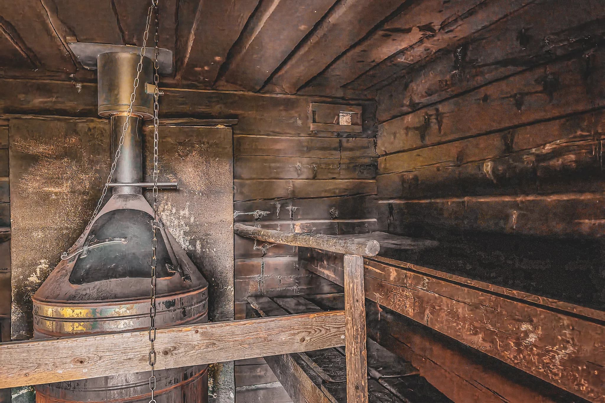 The image shows a rustic wooden interior, featuring a metal stove in the centre. The stove has a conical shape with a flue rising to the ceiling. Metal chains and wooden supports accentuate the stove.