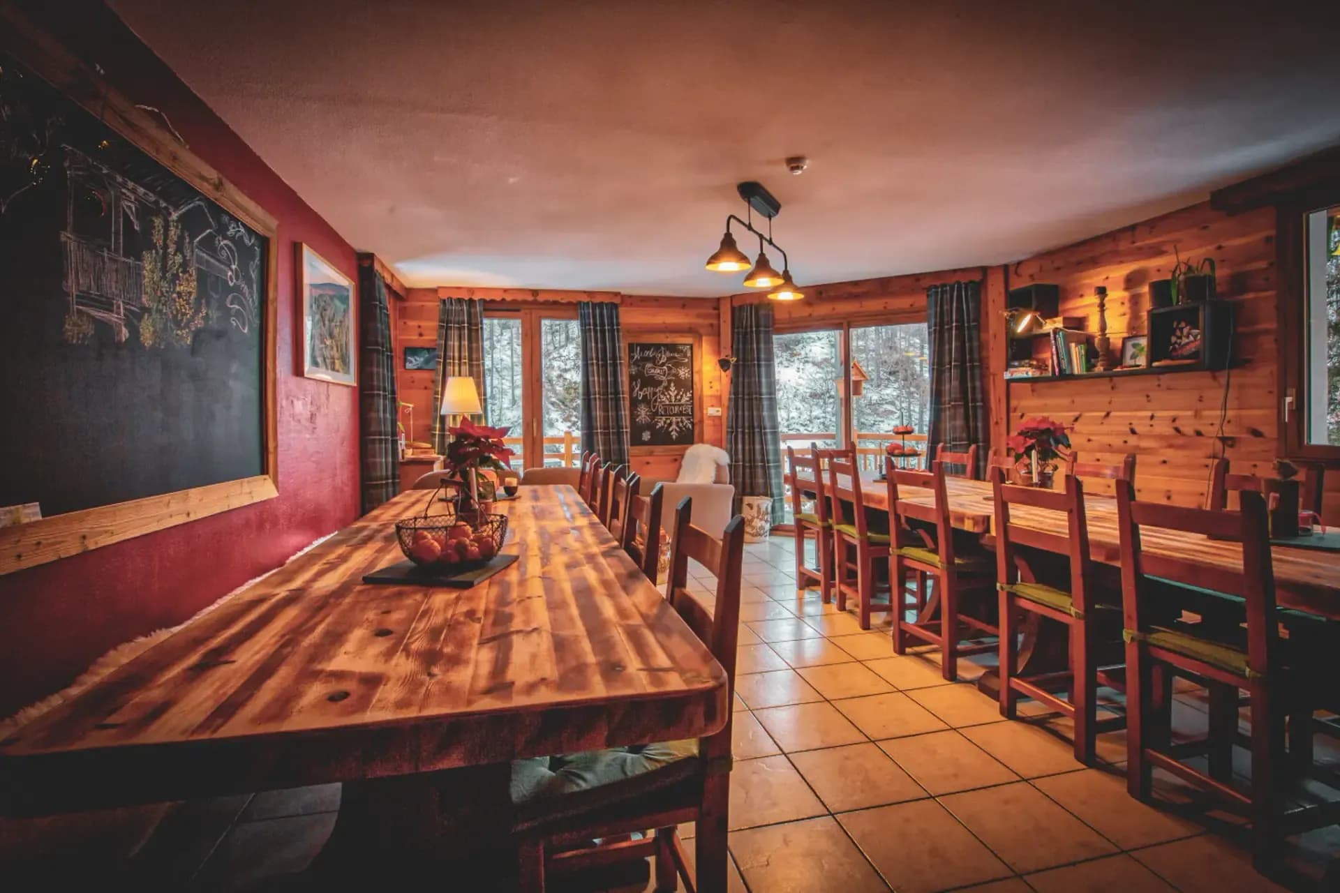 Un espace intérieur chaleureux et accueillant avec de grandes tables en bois, entourées de chaises en bois. Les murs sont peints dans des tons de rouge et les éléments en bois créent une ambiance rustique. Une grande fenêtre laisse entrer la