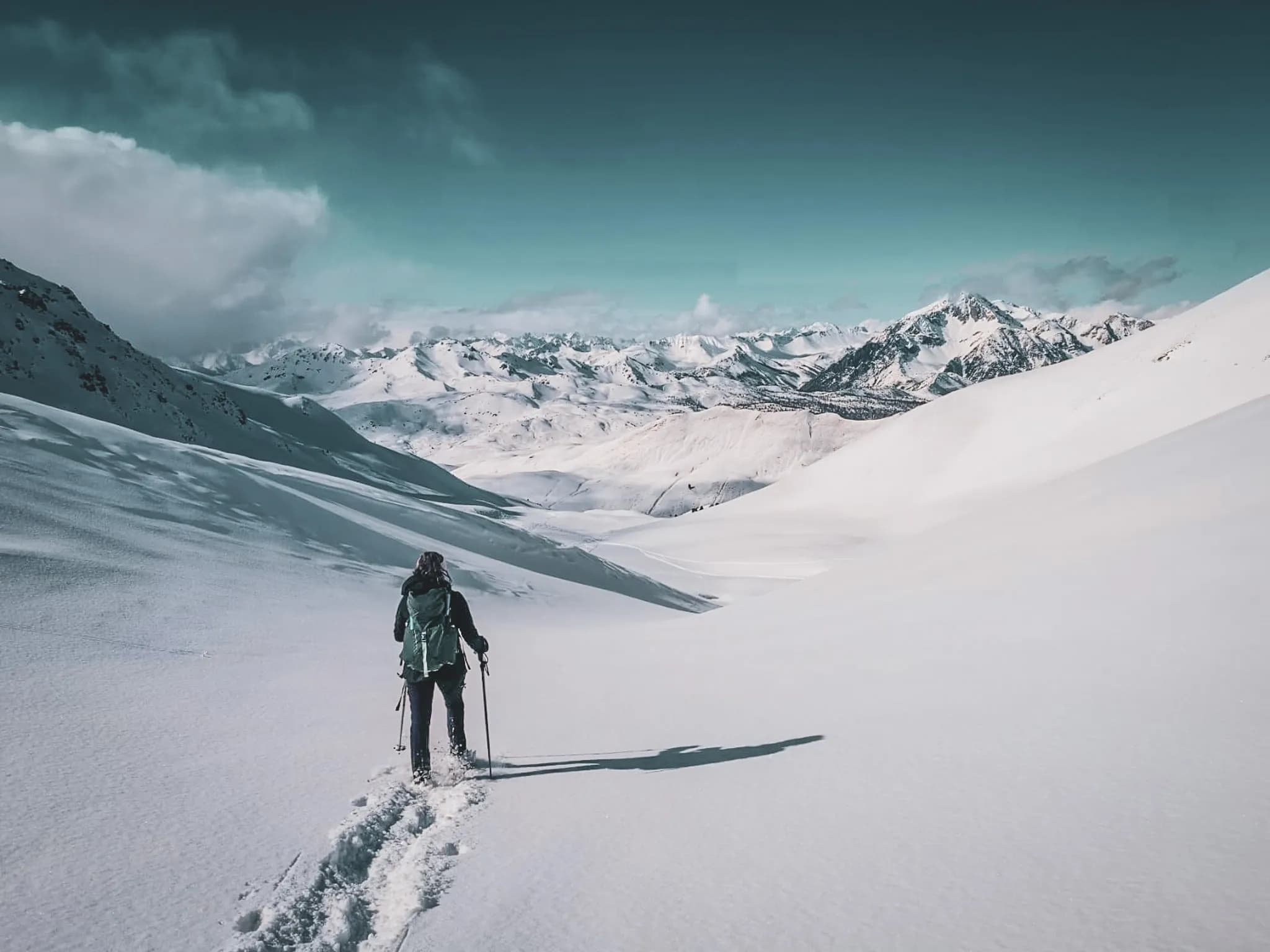 A snowy landscape with two hikers walking on the snow, carrying rucksacks and hiking poles. They are walking along a path cut through the snow, with majestic mountains behind them.