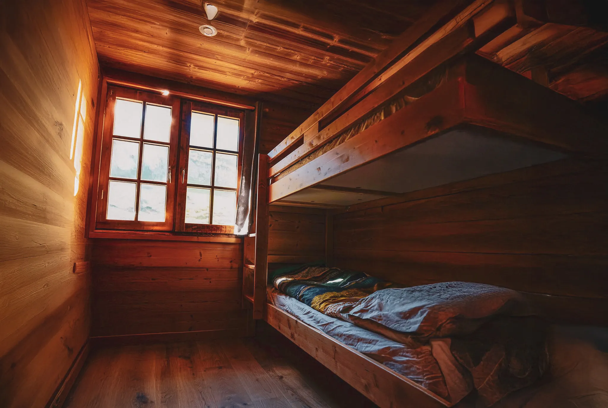 Une chambre en bois chaleureuse et accueillante, dotée de lits superposés en bois. La lumière naturelle pénètre par une grande fenêtre à mulots, illuminant les murs en bois et le sol en bois clair. Sur les lits,
